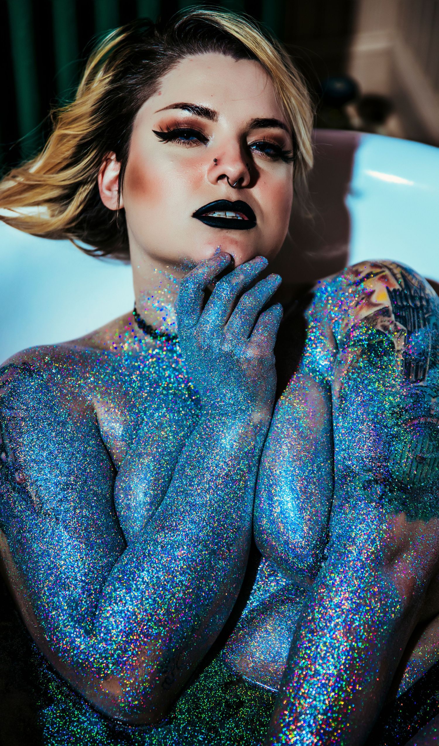 Model covered in iridescent blue glitter body art poses dramatically with dark lipstick in moody lighting.