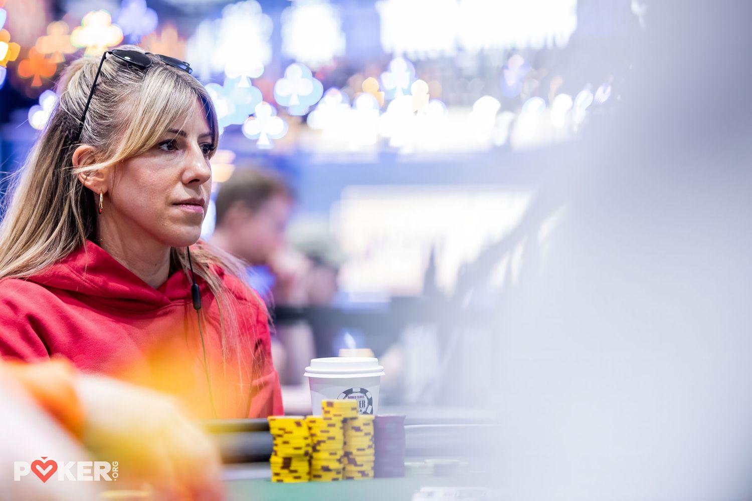 Woman in red jacket focused at a poker tournament table with colorful chips stacked in front of her.