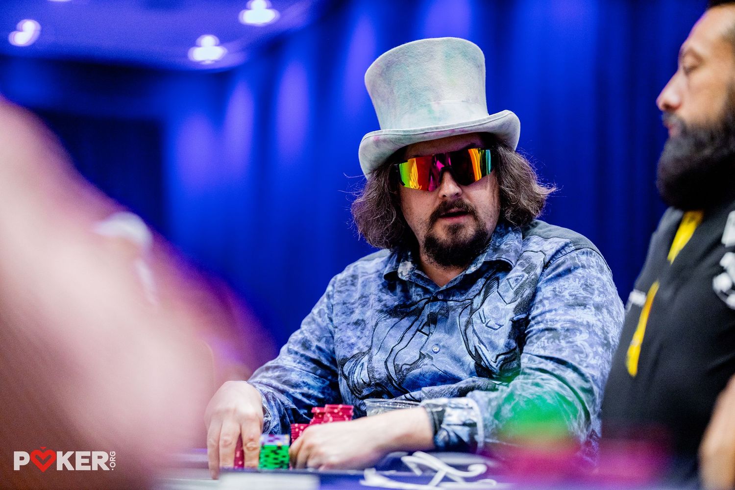 Man in gray top hat and sunglasses playing poker at a casino table with colorful chips stacked in front.