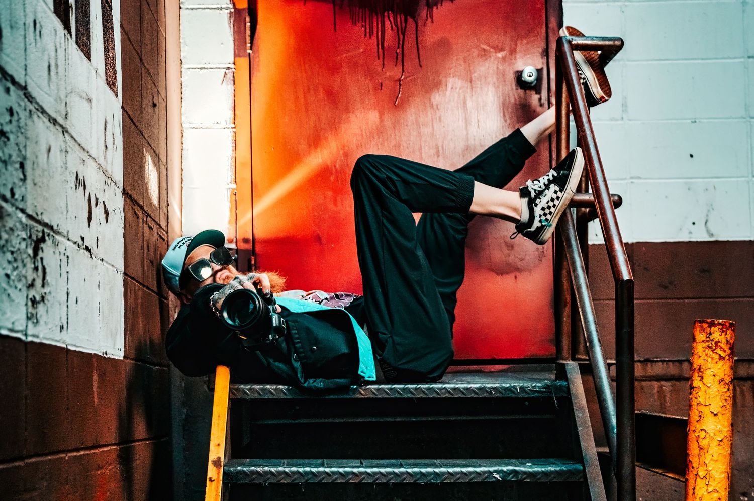 A person in casual wear lies on industrial stairs against a vibrant red wall with dramatic lighting and shadows.