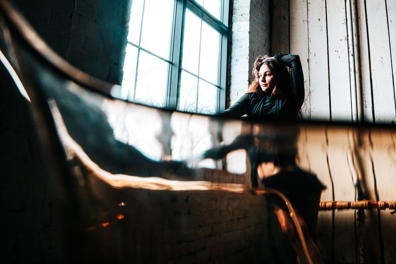 A young woman sits pensively by a large window in a rustic industrial space, soft light illuminating her thoughtful expression.