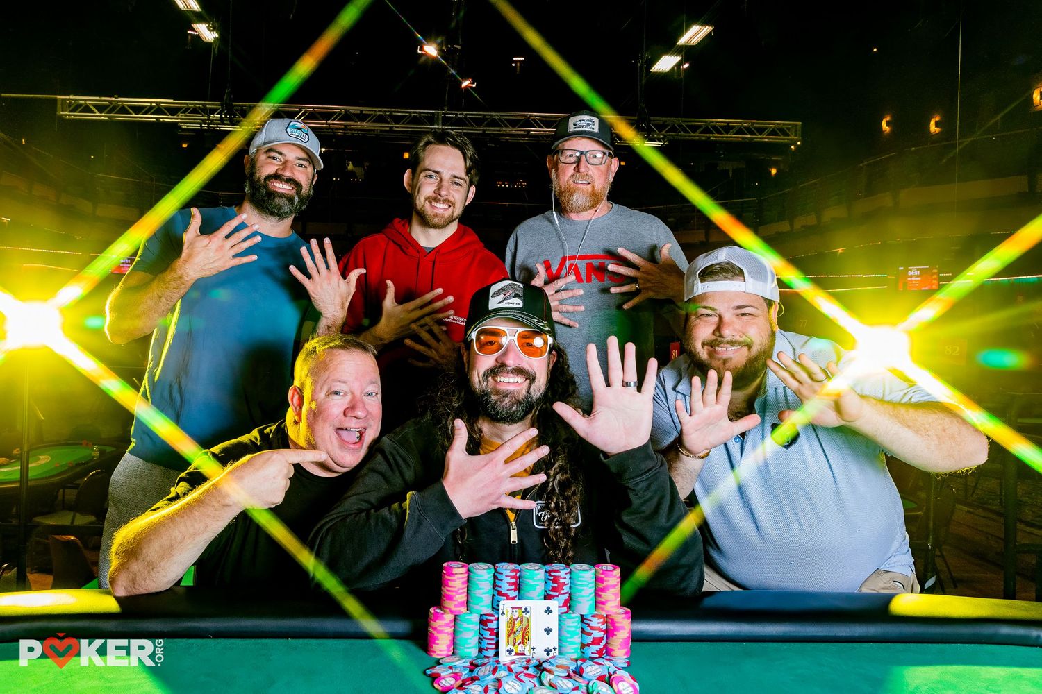 Six poker players celebrating a tournament win with a large chip stack at a WSOP poker event under yellow stage lights.