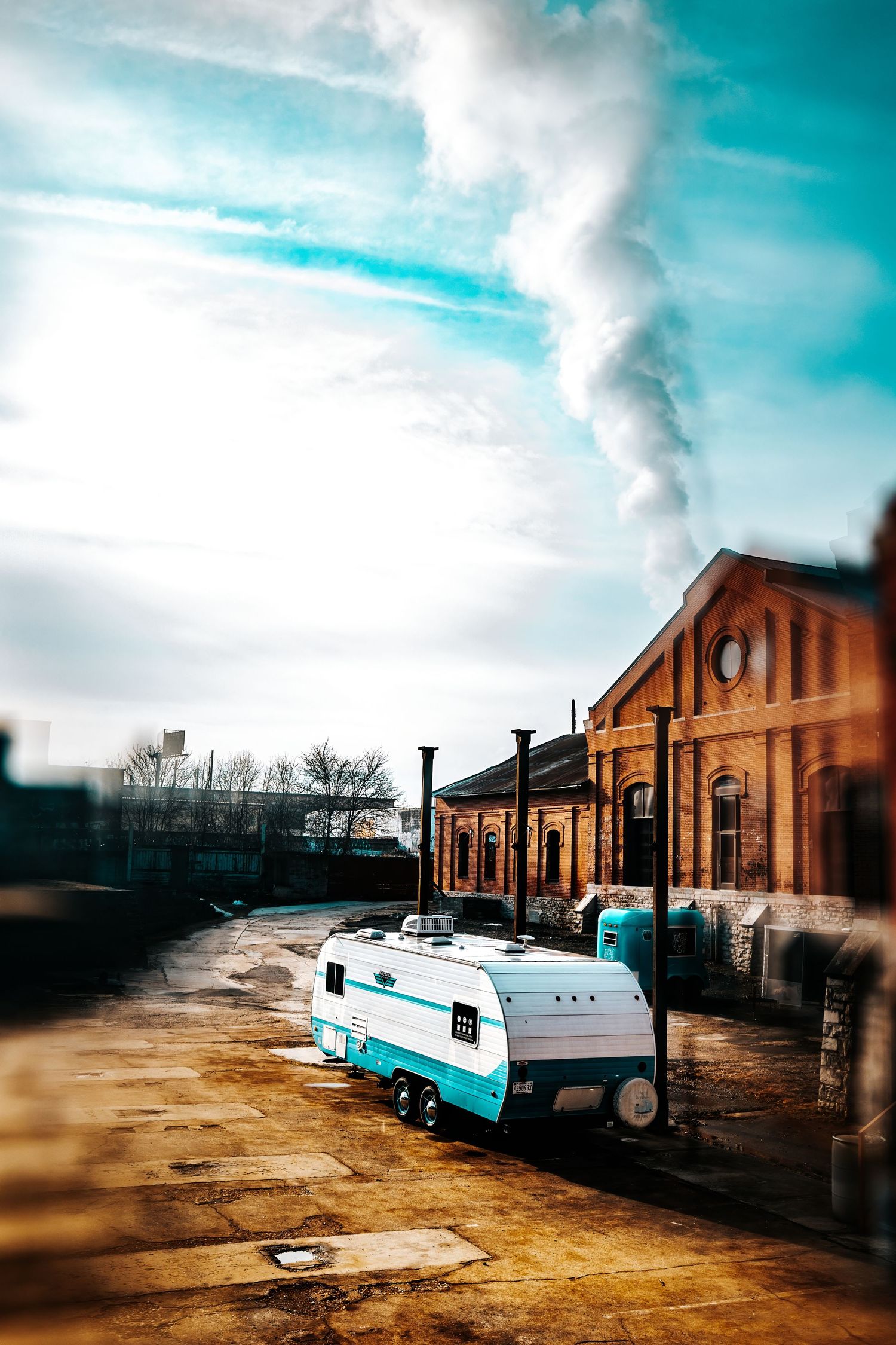 Vintage teal trailer parked beside a historic brick building under dramatic cloudy sky.