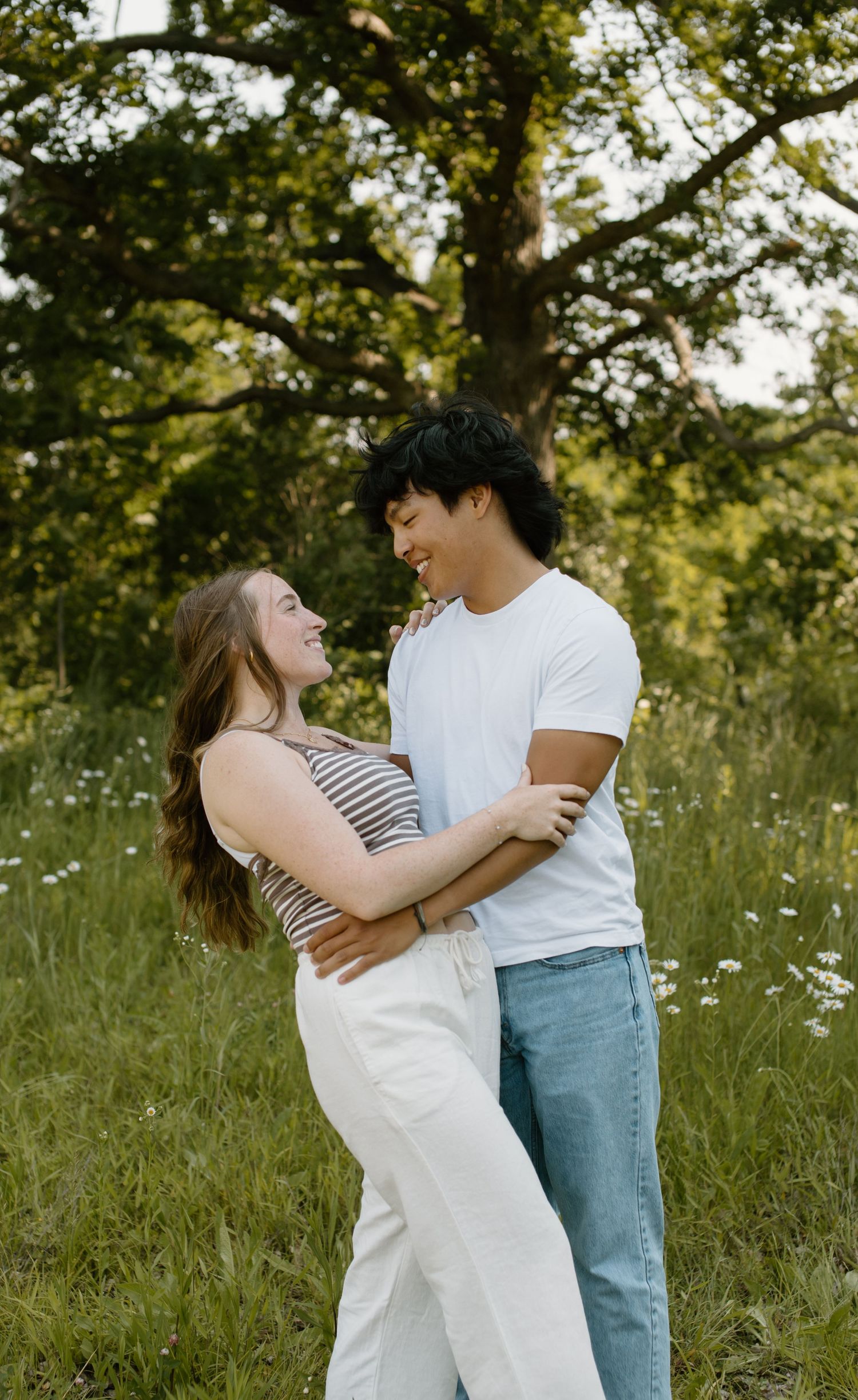 A couple embraces in a sunny meadow with wildflowers and trees in the background during golden hour.