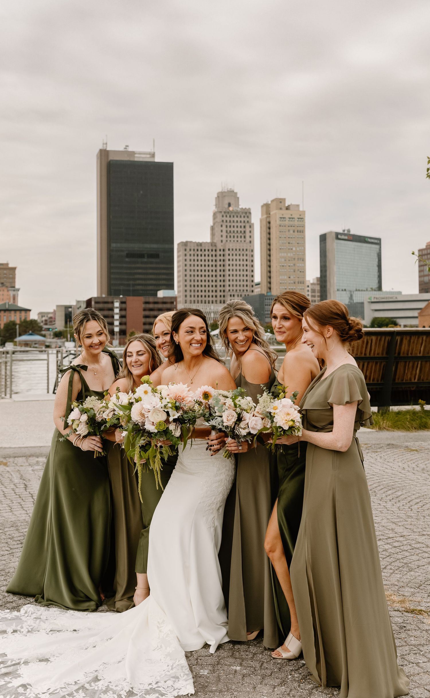 Bridal party in olive green dresses poses together on waterfront with city buildings in background.