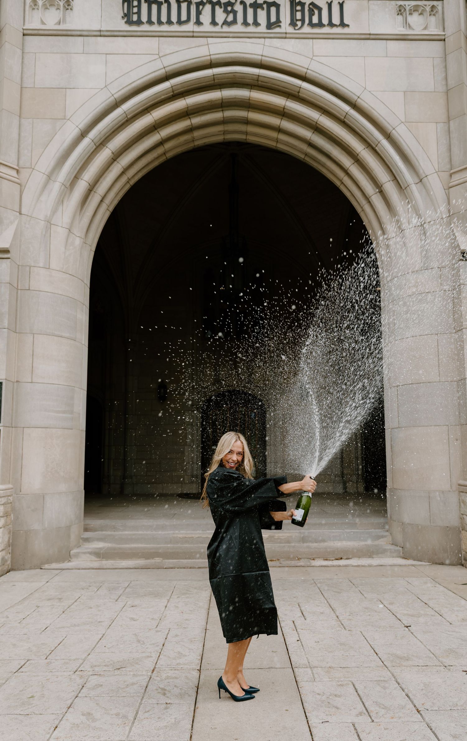 Woman in black dress spraying champagne bottle in front of University Hall stone archway.