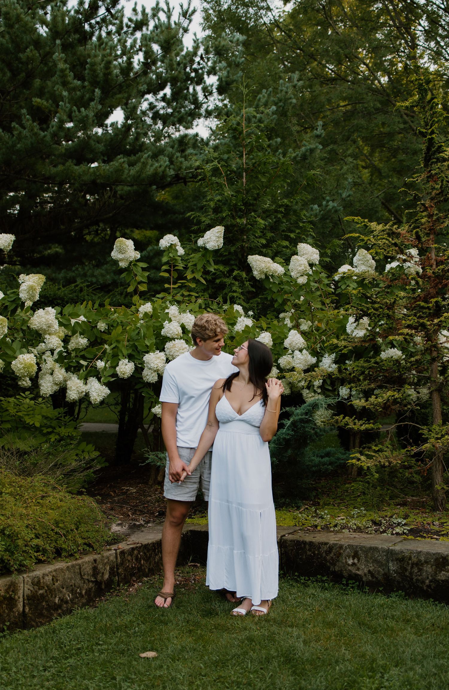 A couple holds hands and gazes at each other in a lush garden with white hydrangeas blooming behind them.