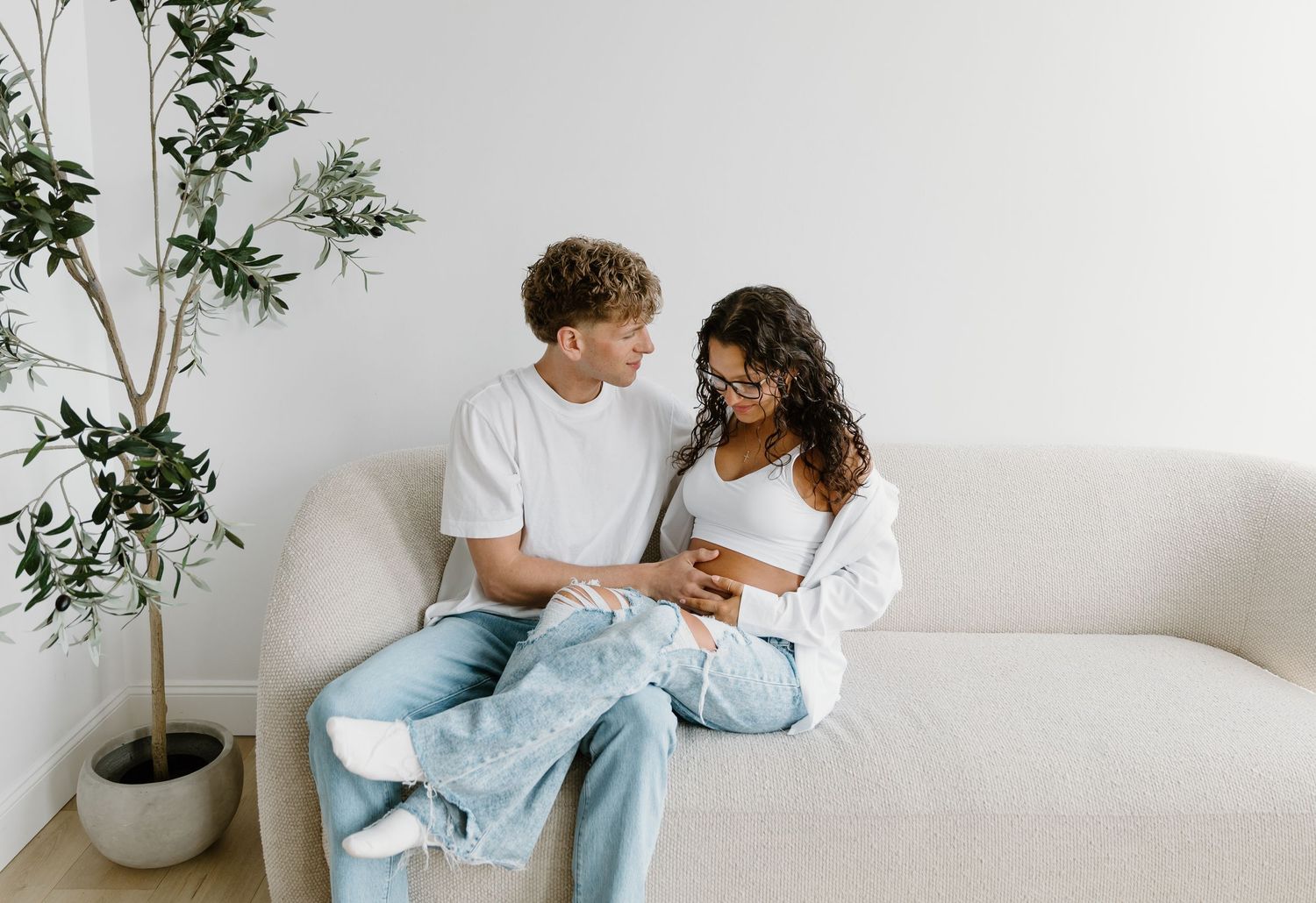 A young couple sits closely on a white couch, smiling at each other in a bright modern living room with a potted plant nearby.