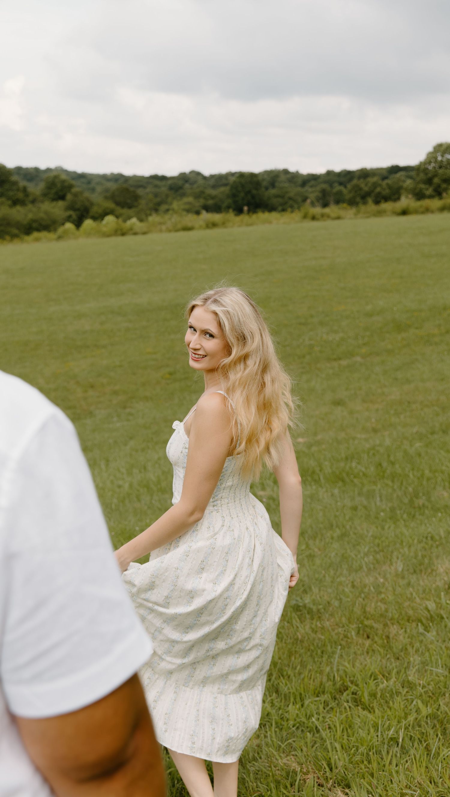 A girl wearing a flowy white summer dress twirls in a scenic green meadow in cuyahoga falls on a cloudy day.