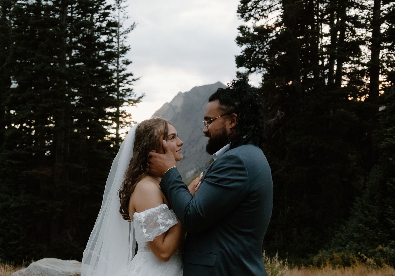 A couple embraces in a romantic outdoor setting with evergreen trees silhouetted against the sky at dusk.