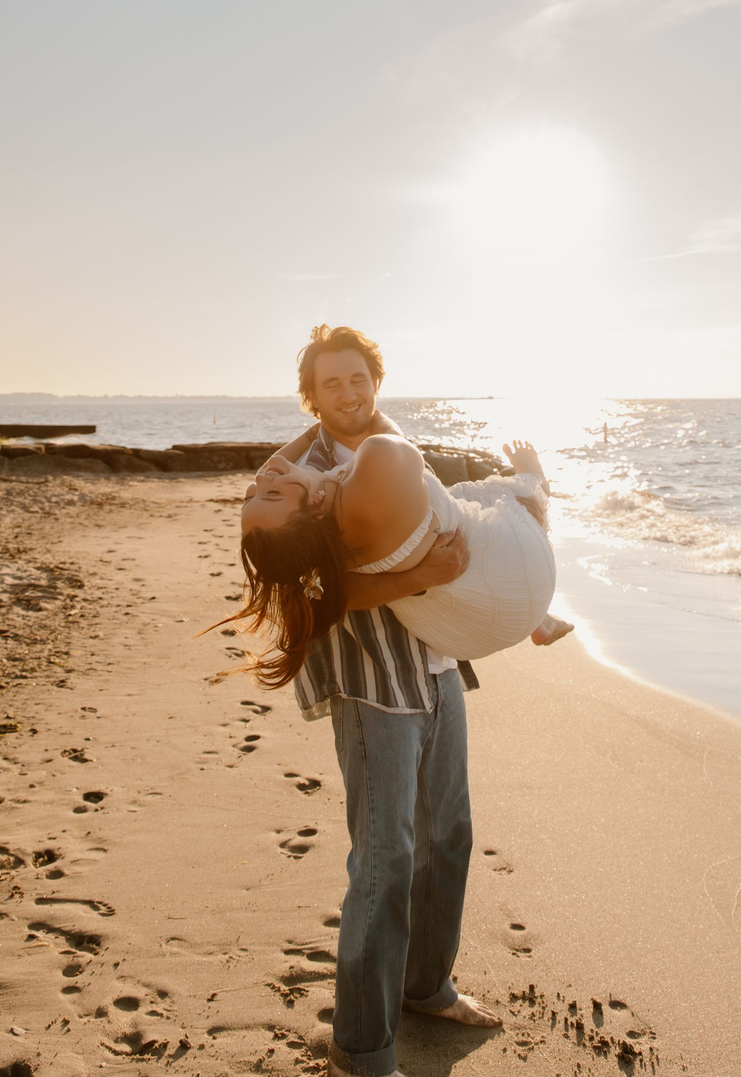Couple laughing on a sunny beach at golden hour, man lifting woman in his arms near the ocean waves.