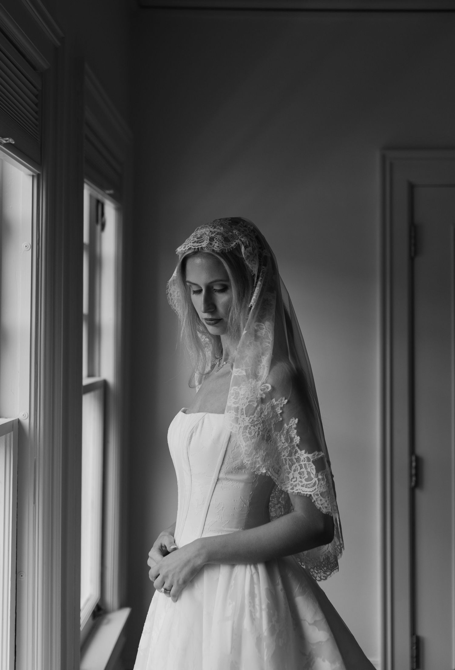Black and white portrait of bride in lace veil and strapless gown standing by window in soft light.