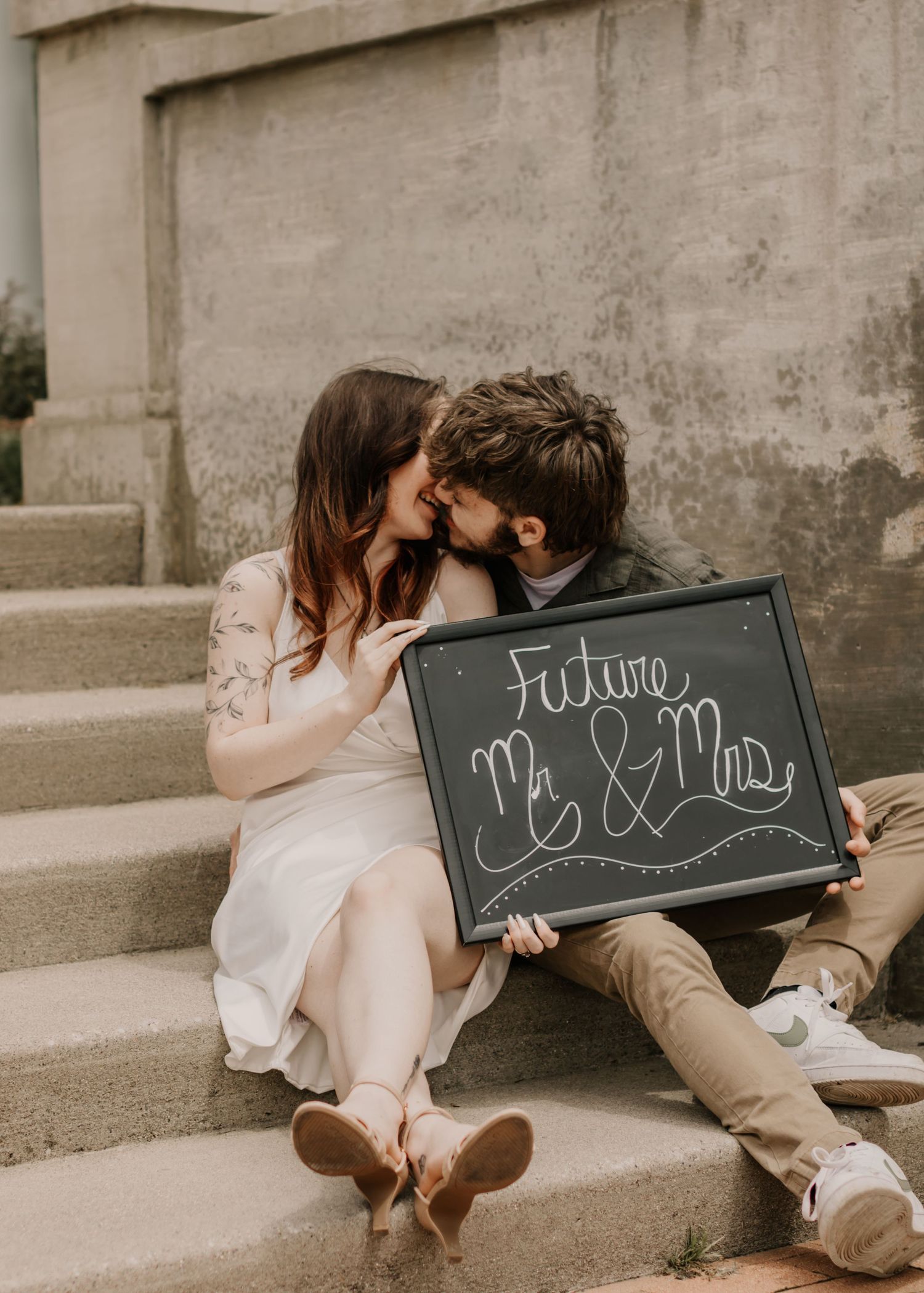 A couple shares an intimate moment on concrete steps while holding a chalkboard sign.