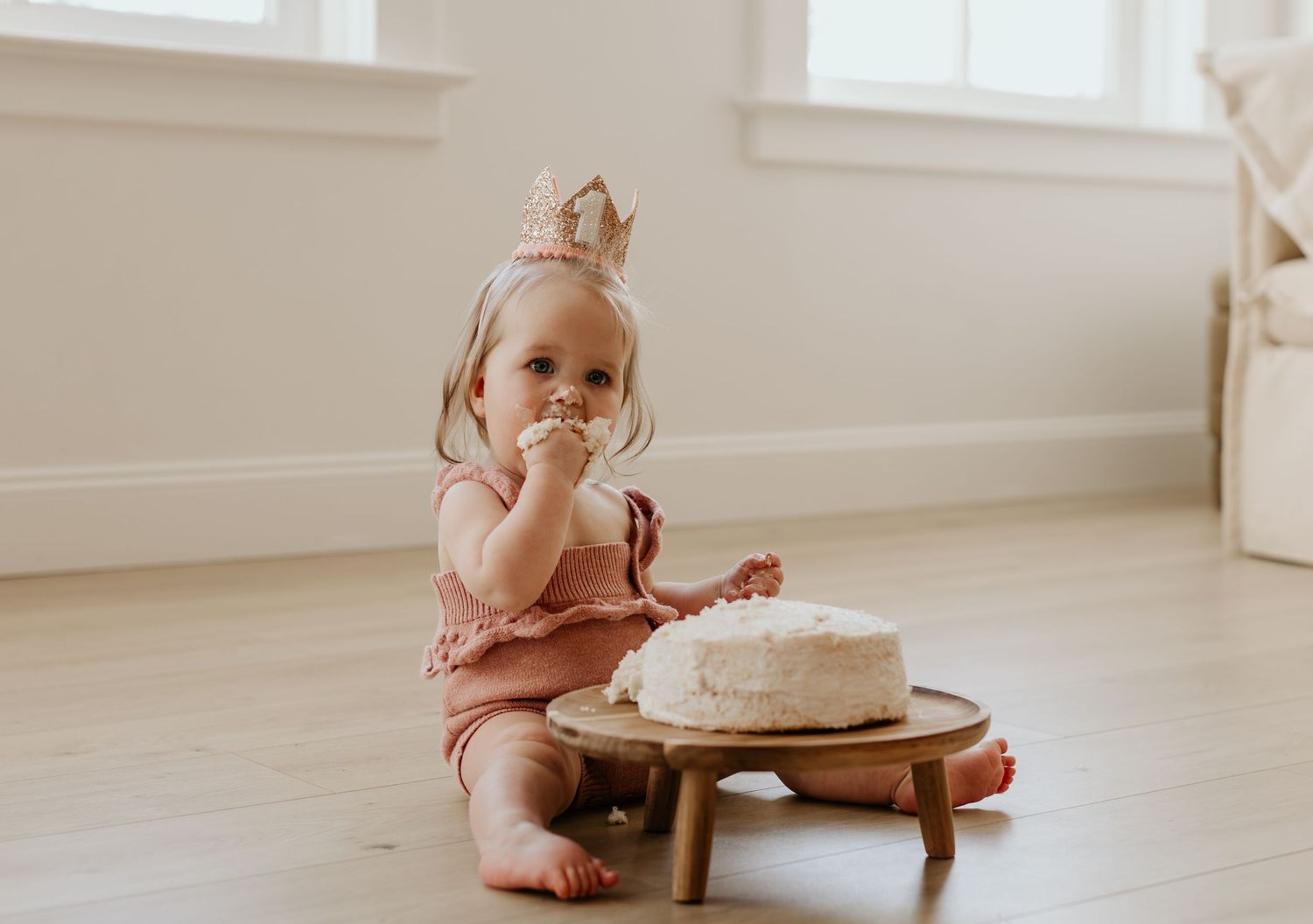 Small child in pink outfit enjoying birthday cake on wooden surface.