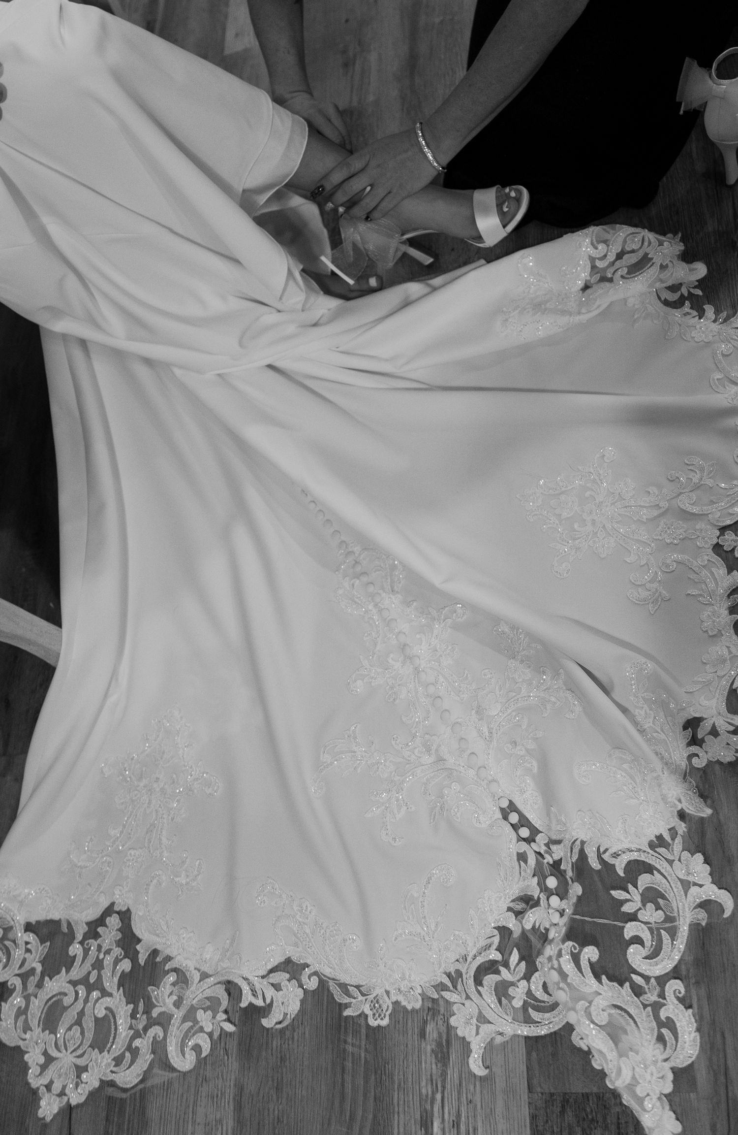 Elegant lace-trimmed wedding dress train spread on wooden floor with bride and groom shoes visible above.
