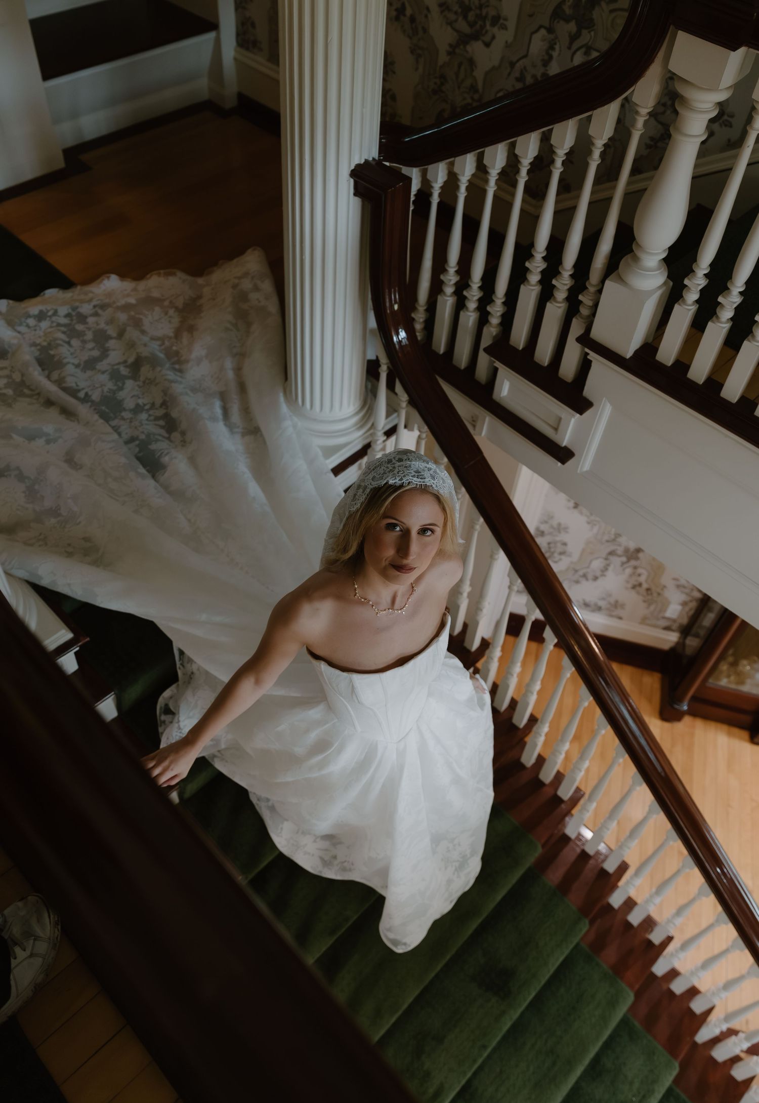 Bride in white wedding gown and tiara looks up from grand staircase with green carpet and white columns.