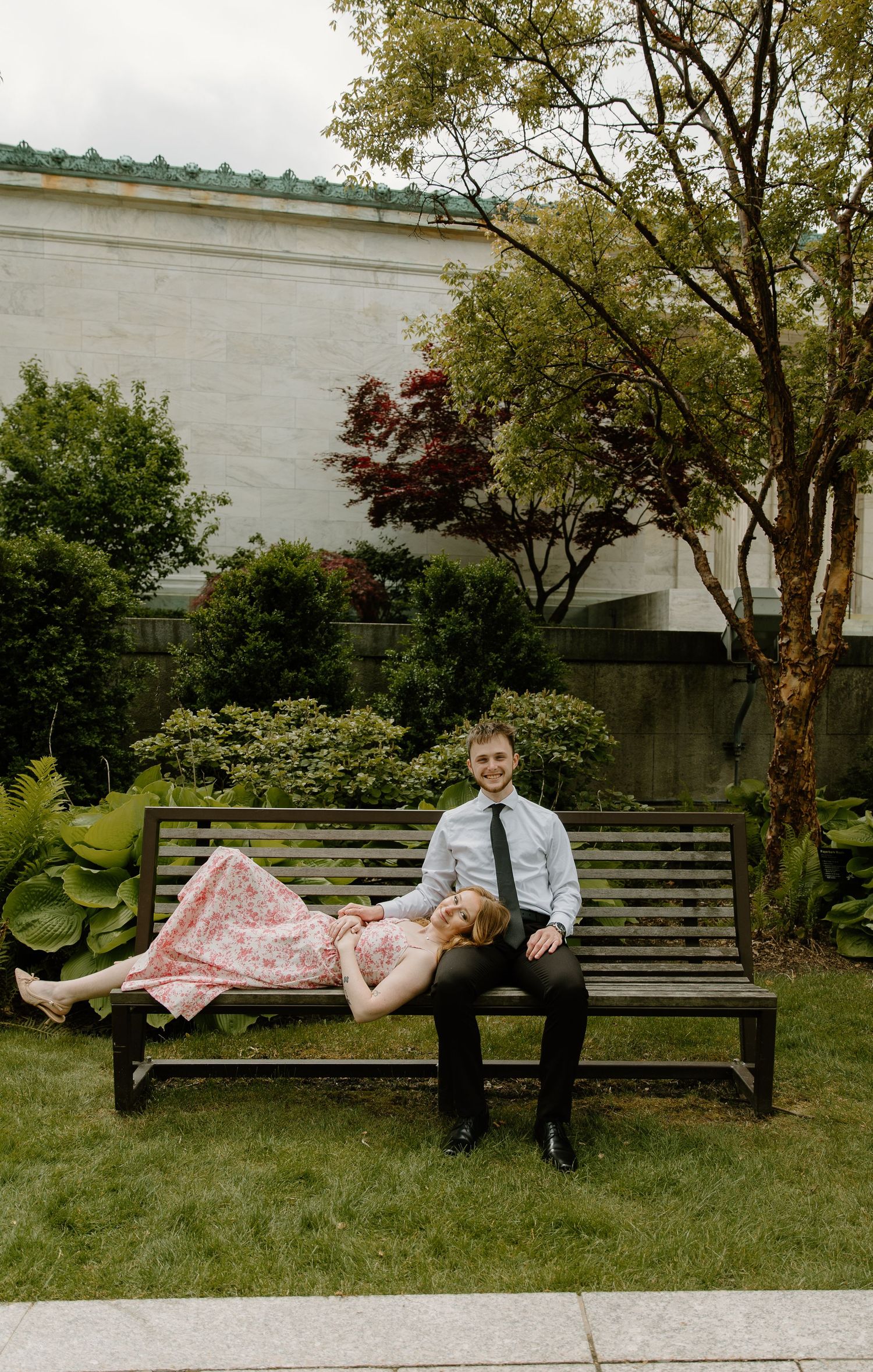 Couple posing on a wooden bench in a garden, woman lying across man's lap in a pink floral dress.