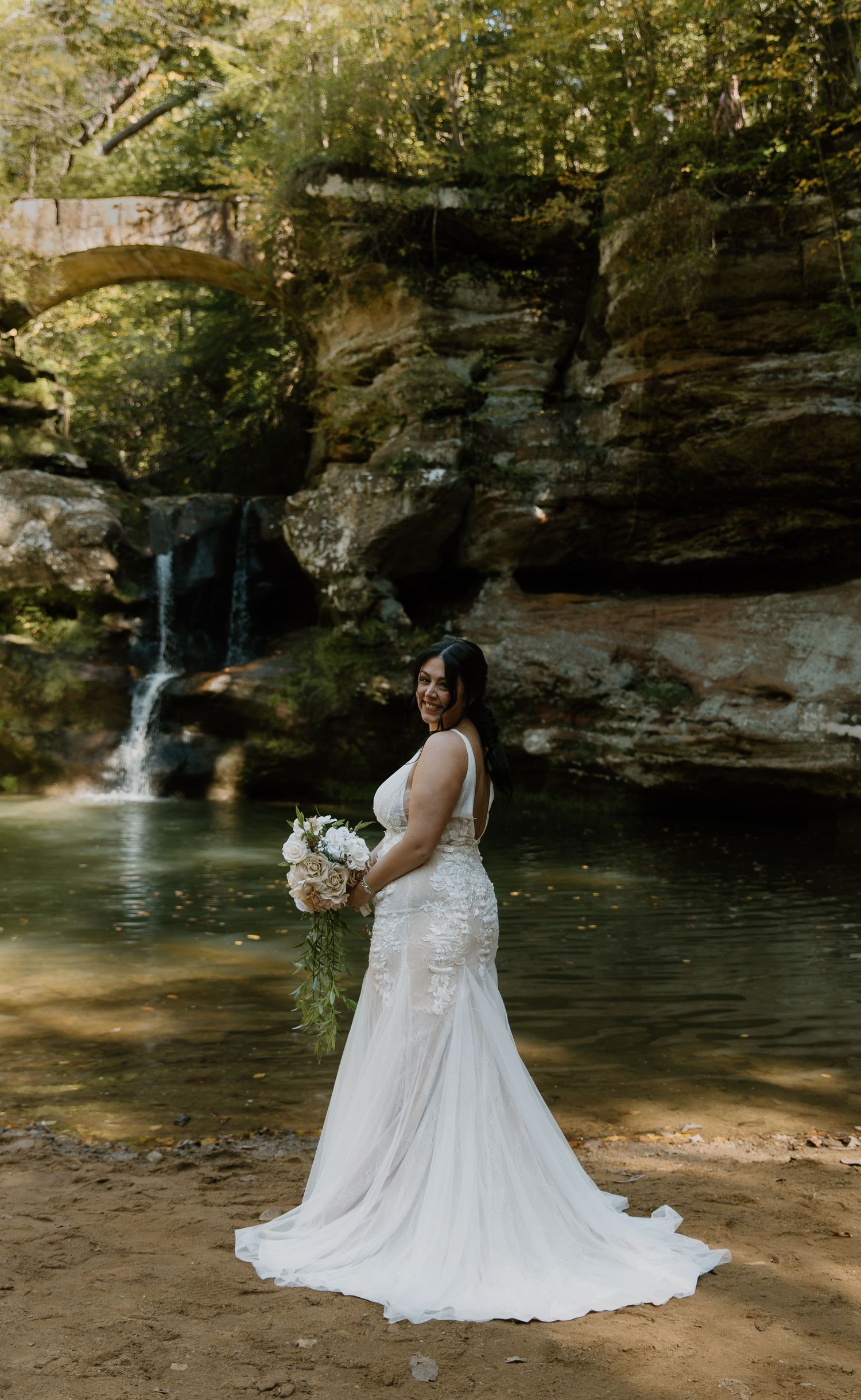 Smiling bride in fitted lace gown poses by waterfall and stone arch bridge in Hocking Hills, Ohio.