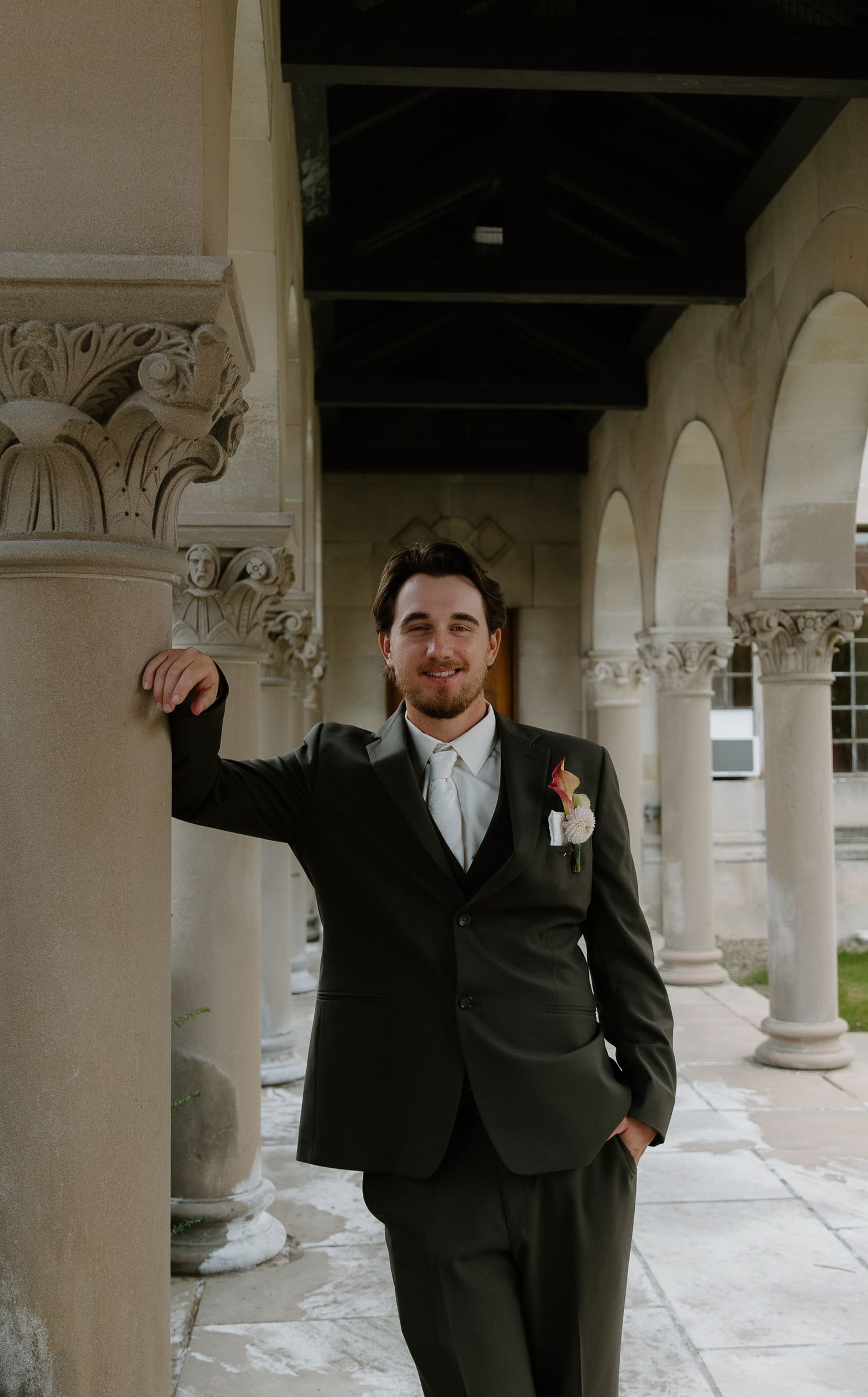 Groom in dark suit with boutonniere leans against stone column in elegant arched colonnade.