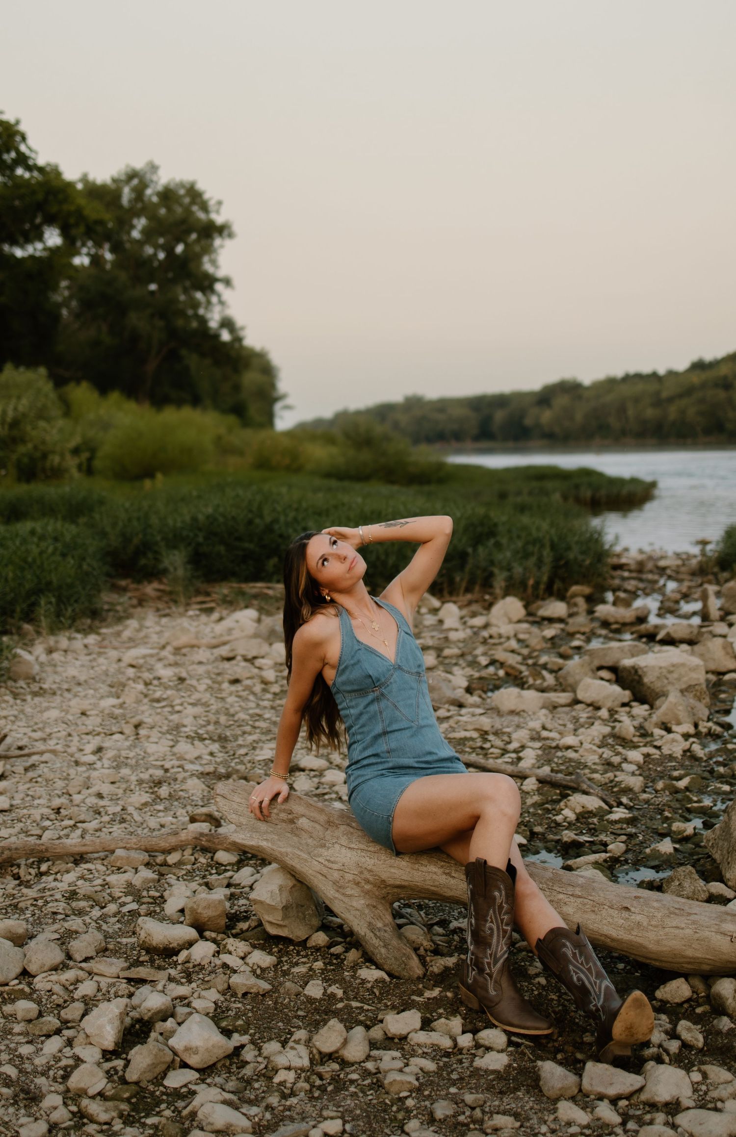 Woman in blue dress and cowboy boots sitting on driftwood by a rocky riverbank at golden hour.