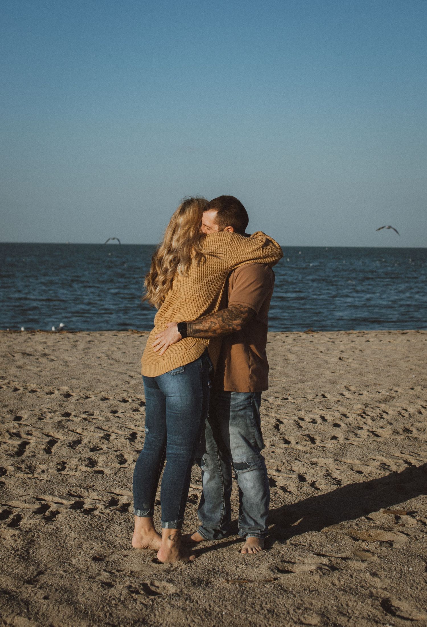 Couple in matching brown sweaters hugging on beach with seabirds flying overhead.