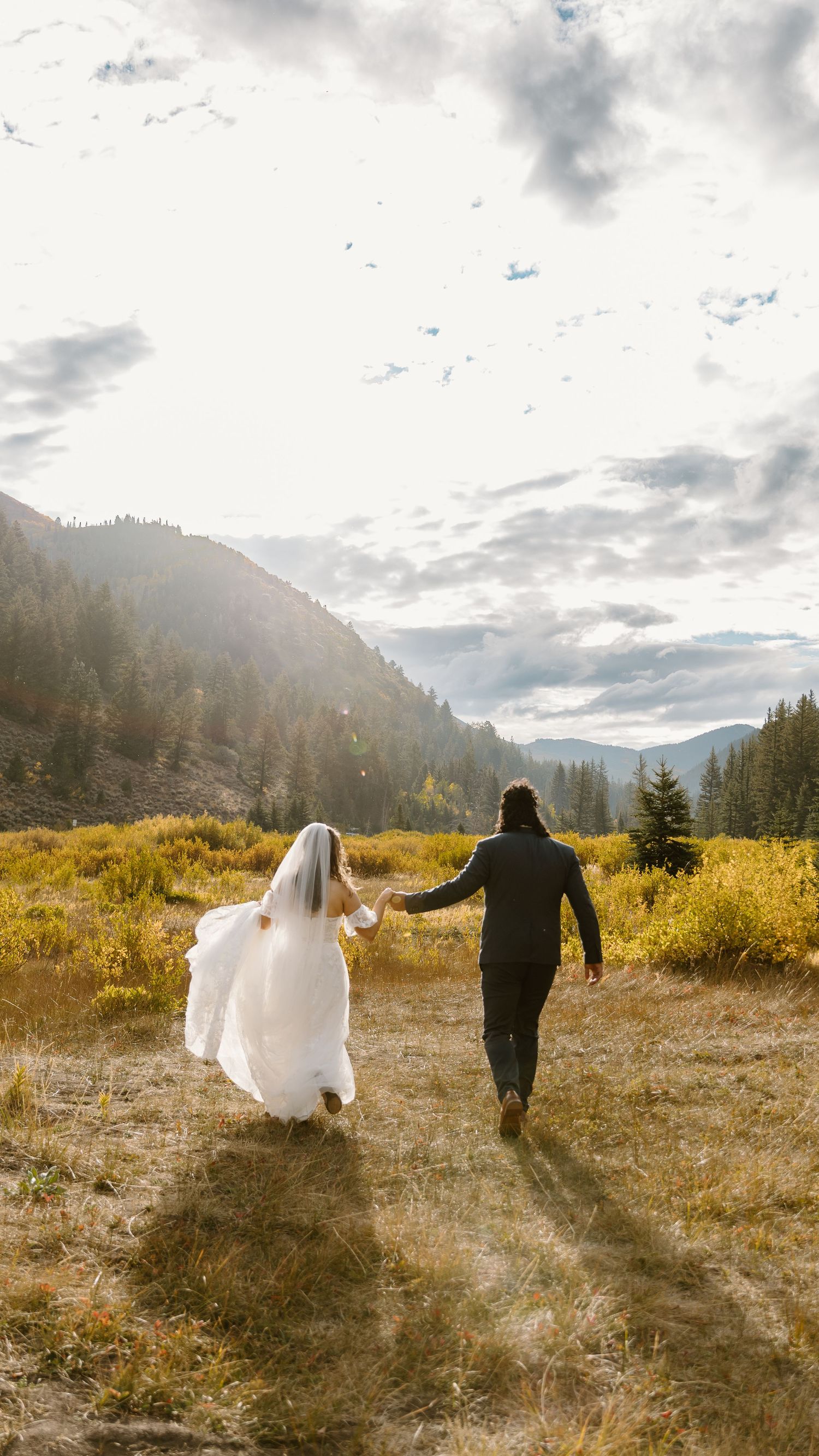 A couple in wedding attire runs through a golden meadow with mountain views and dramatic clouds at sunset.