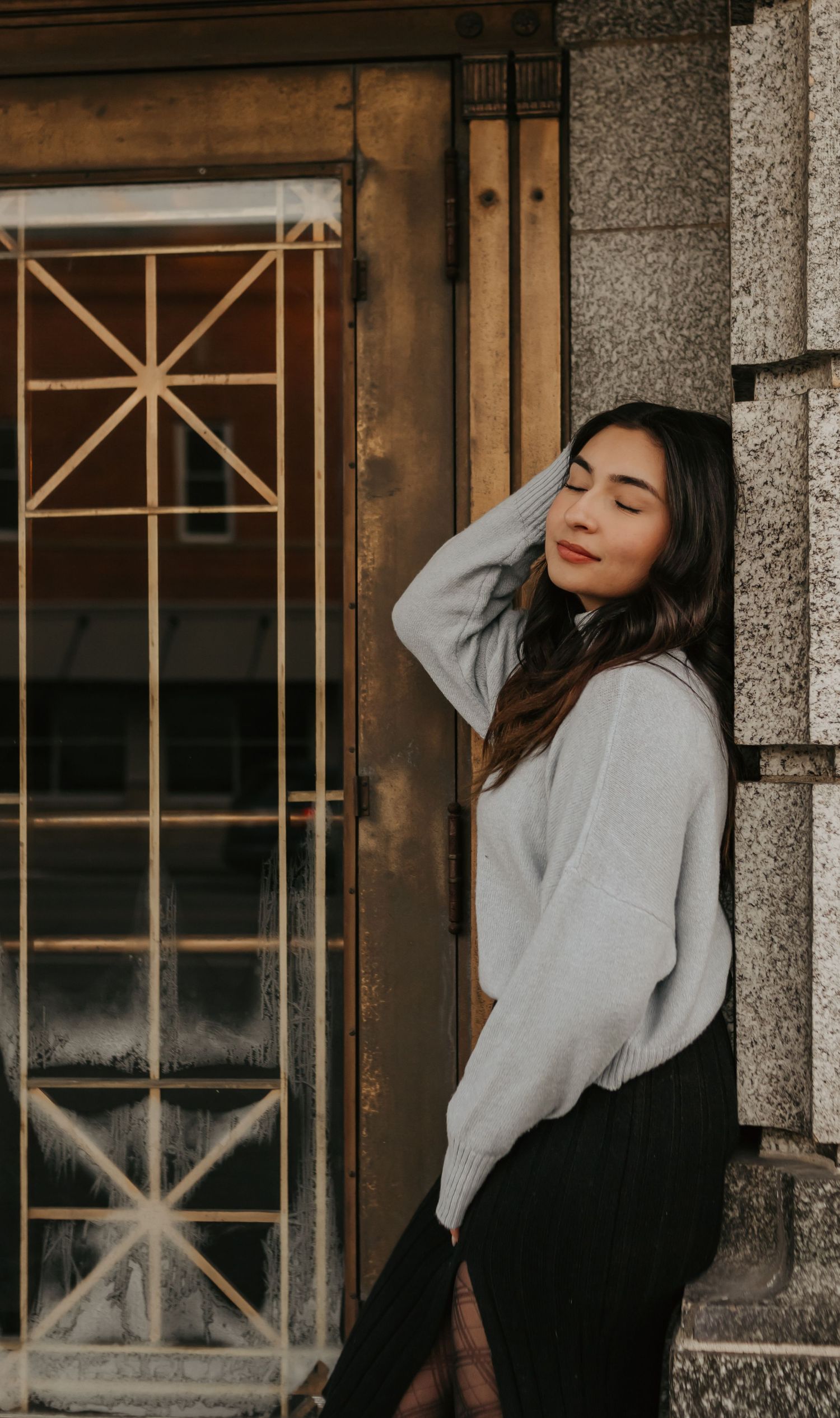 Person in grey sweater poses against stone wall with decorative window.