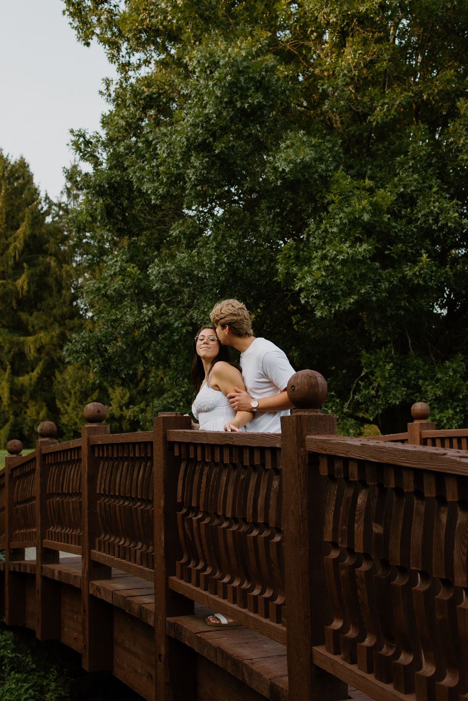 A couple embraces romantically on a ornate wooden bridge surrounded by lush green trees in a garden setting.