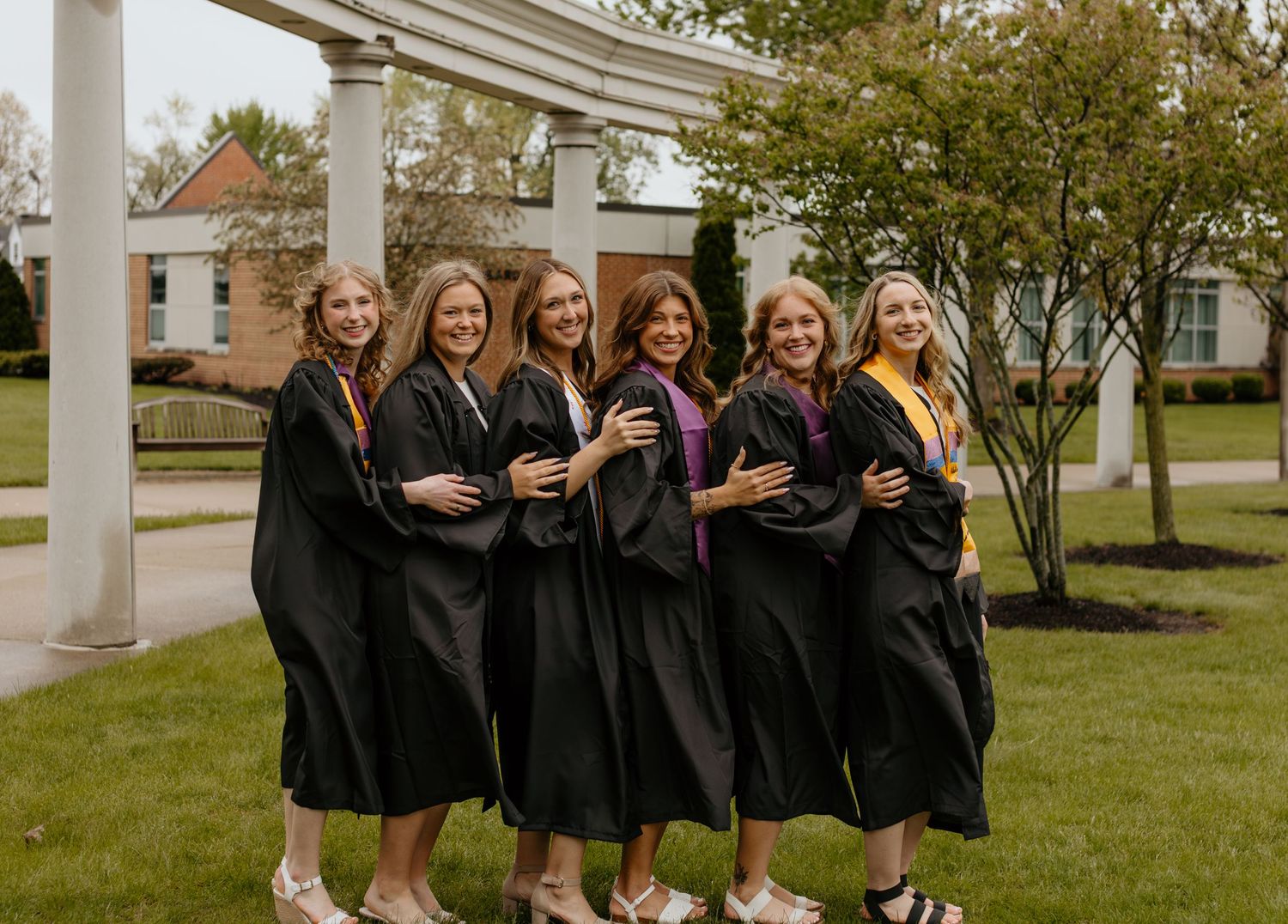 A group of graduates in black robes pose together with drinks under a white pergola on a sunny day.
