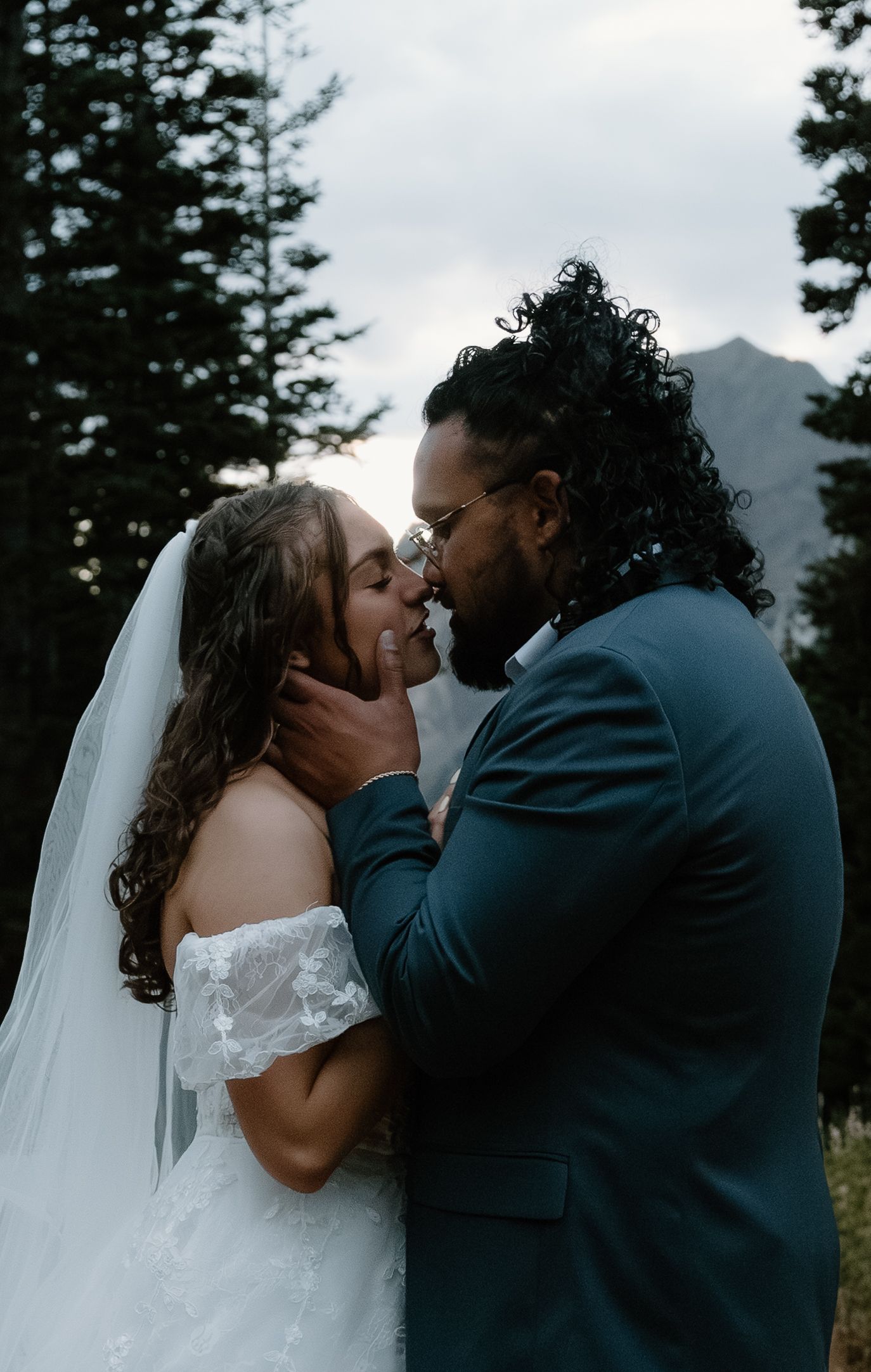 A romantic wedding couple shares a tender kiss against a backdrop of evergreen trees and mountains at dusk.