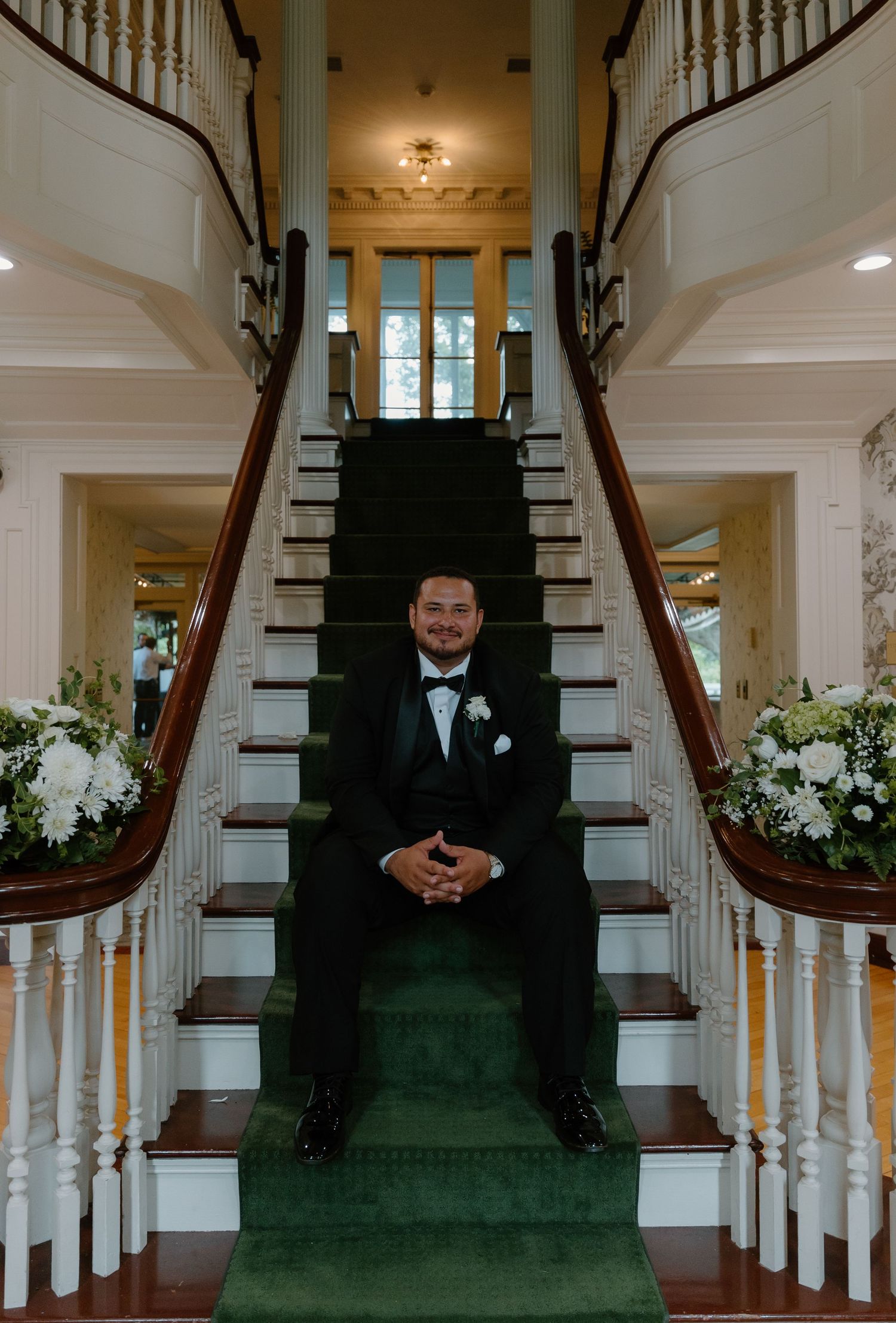 Smiling groom in black tuxedo sits on green-carpeted staircase flanked by white floral arrangements.