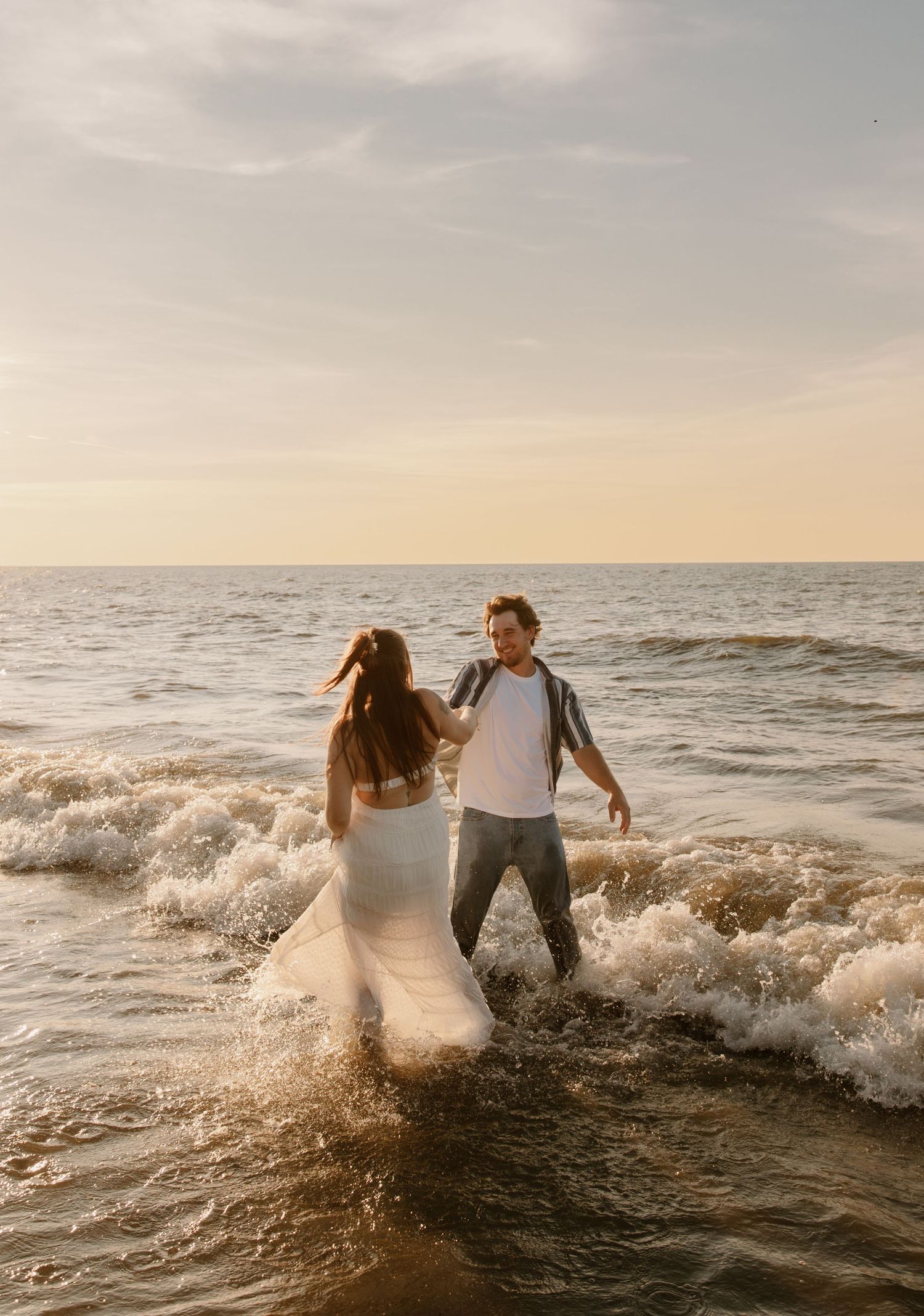 Couple in wedding attire wading through ocean waves at sunset on a beautiful beach.