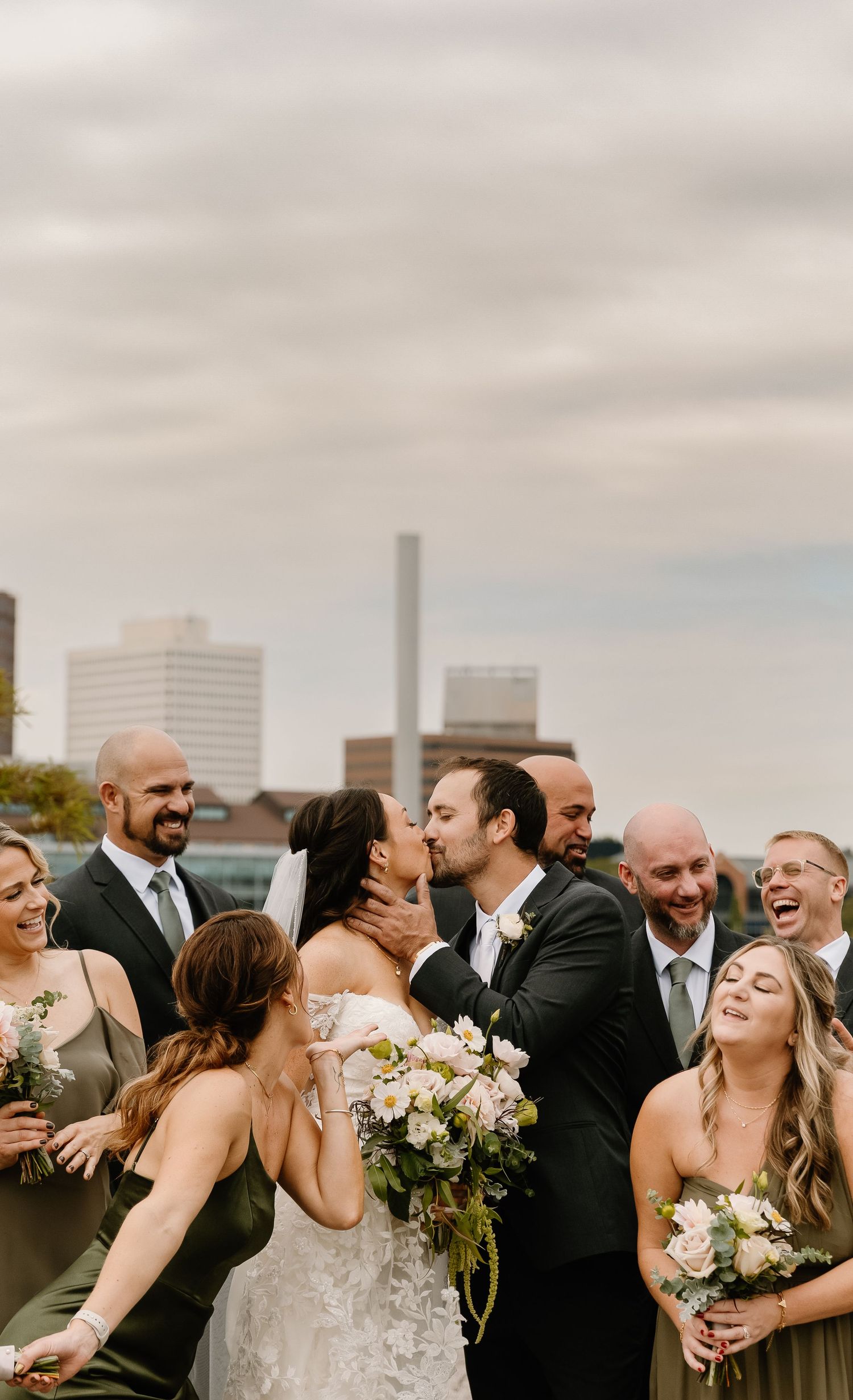 A joyful wedding party laughs and celebrates together on a rooftop with city buildings in the background.
