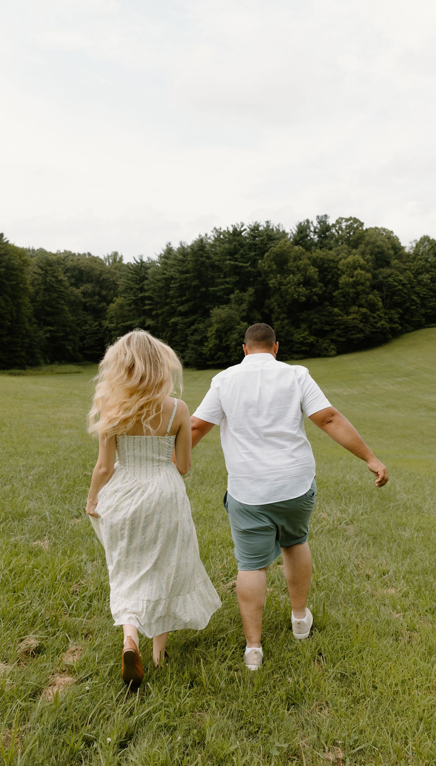 Two people holding hands while running through a lush green cuyahoga meadow wearing white and casual summer clothing.