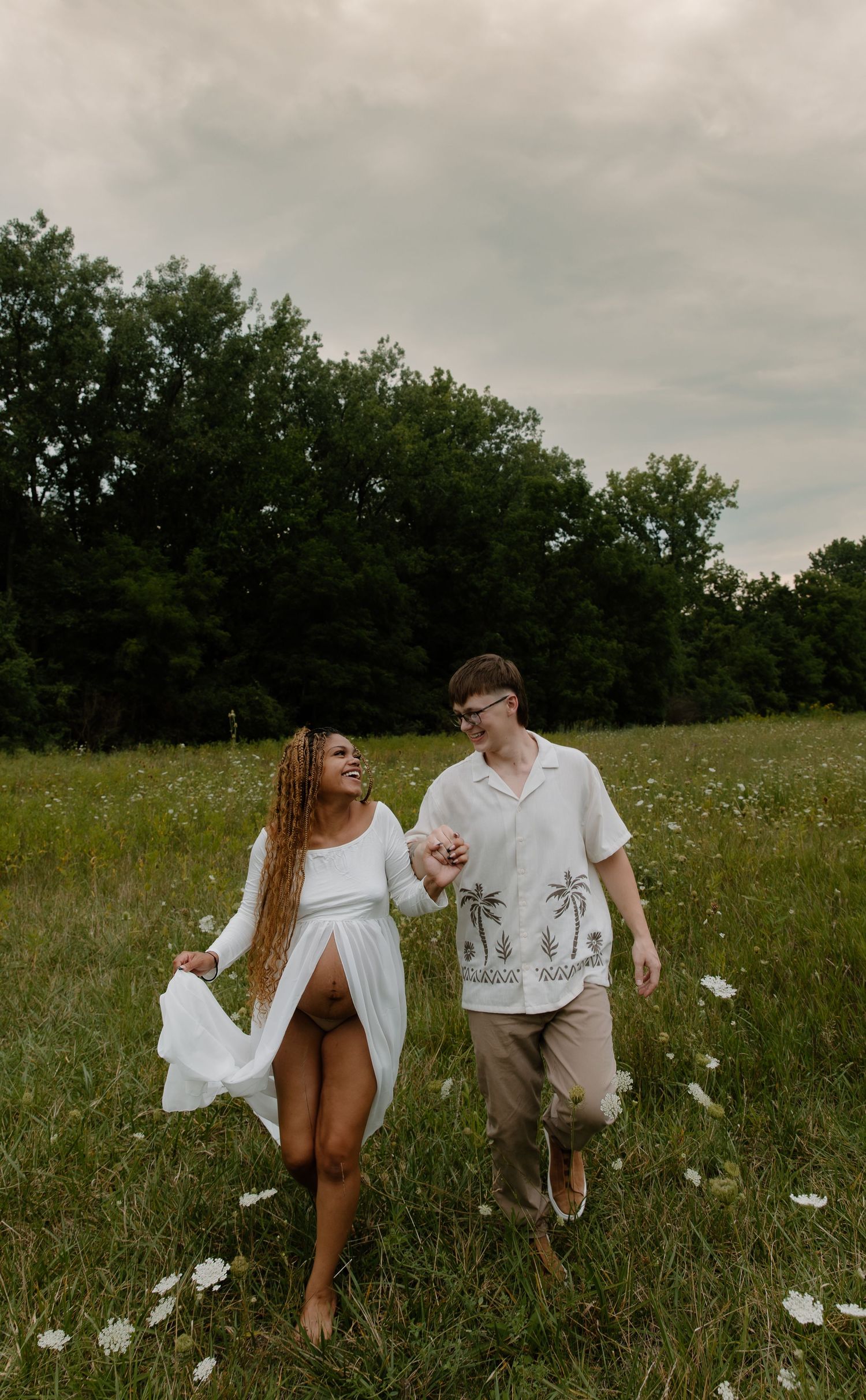 A couple walks hand-in-hand through a wildflower meadow, smiling at each other on a cloudy day.
