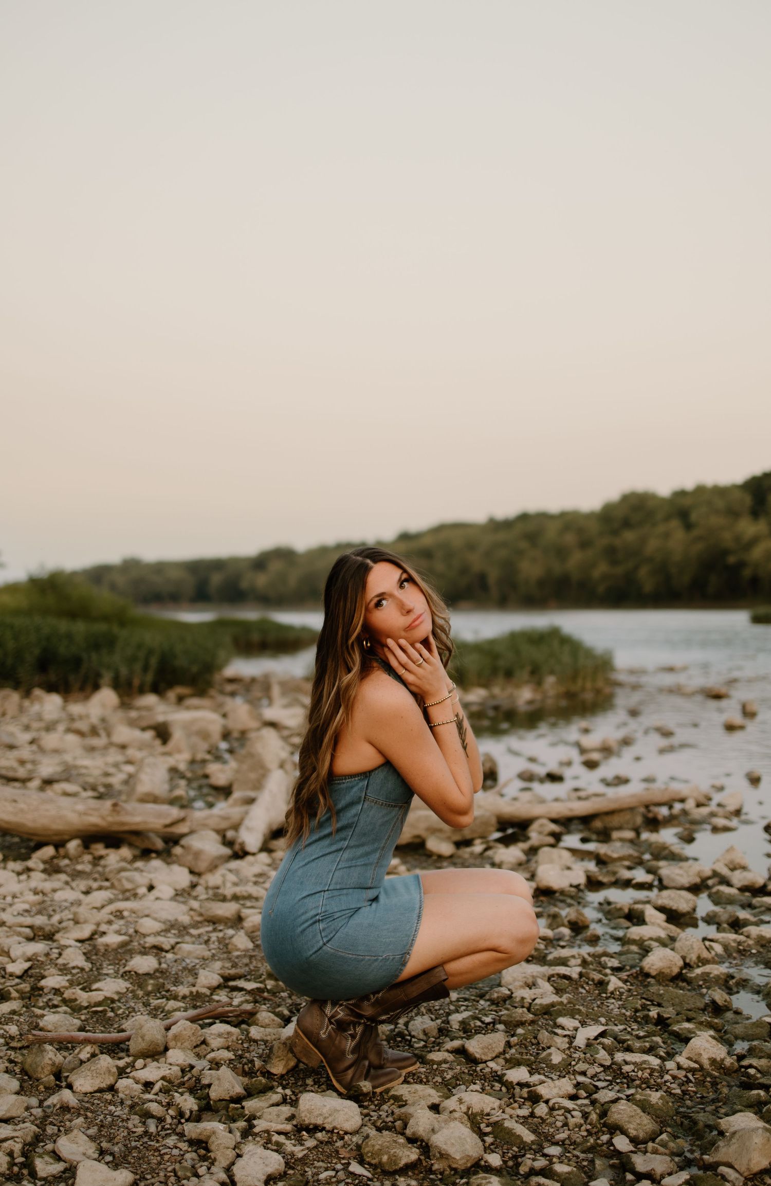 Young woman in blue mini dress posing on rocky riverbank at golden hour with lush greenery in background.