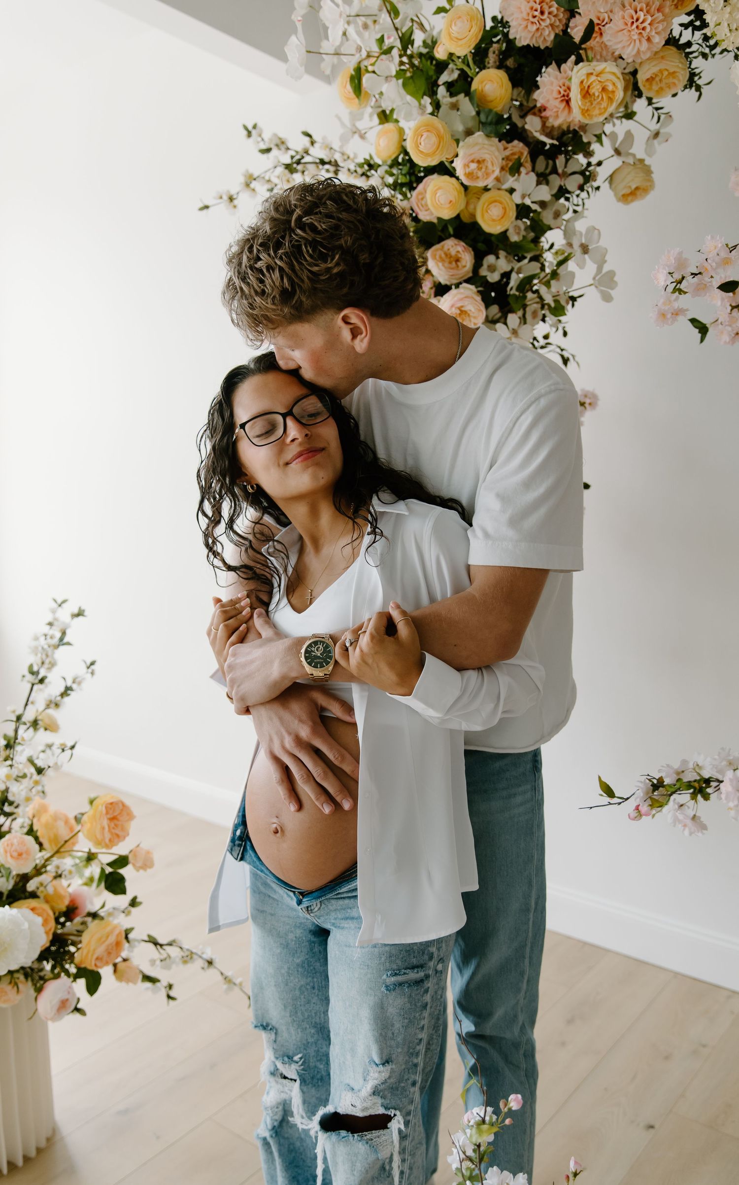 Couple embracing tenderly in front of floral arrangement, pregnant woman with glasses leaning into partner's kiss.