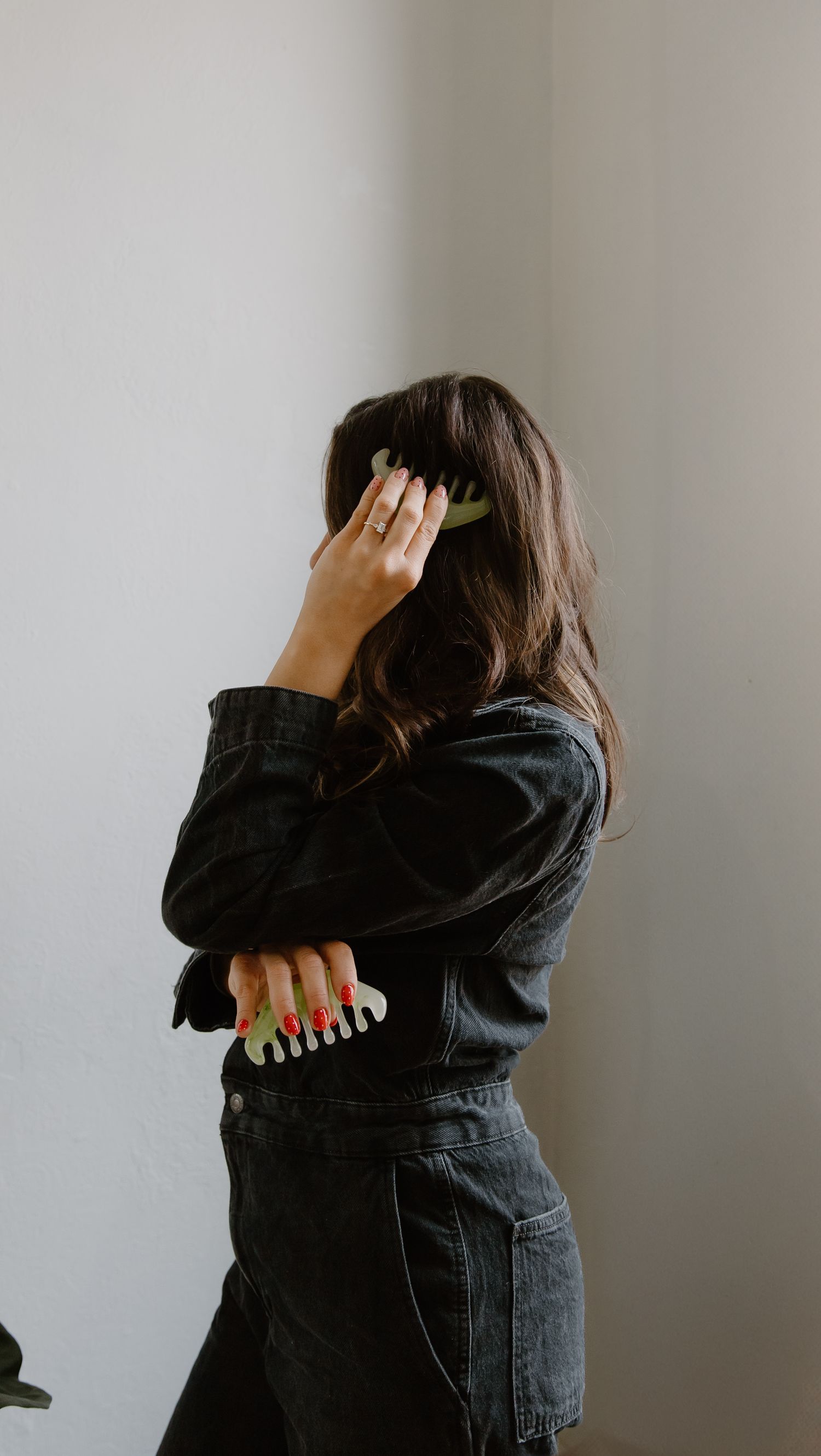 Woman in black denim jacket holding hair clips, covering face with hand against white wall.