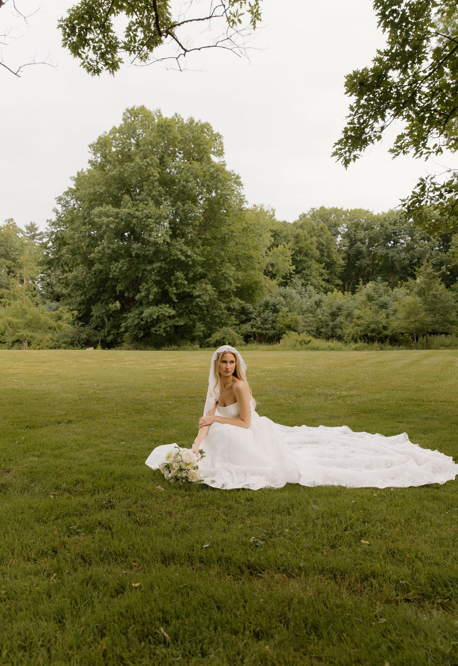 Bride in a lace wedding dress and veil sitting gracefully on a green lawn surrounded by trees.