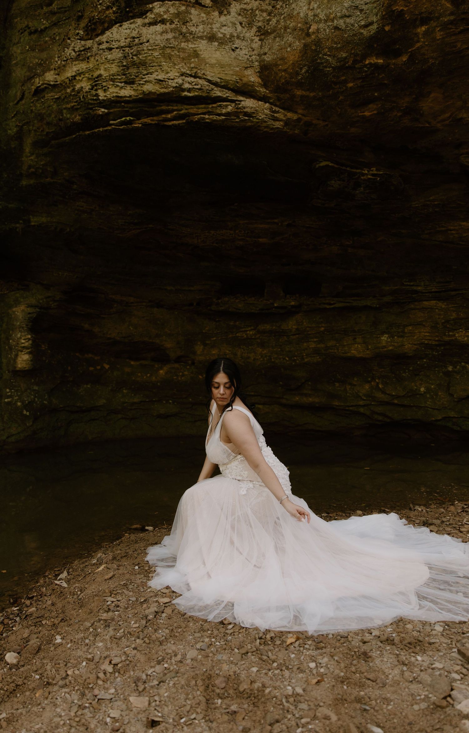 Bride in a flowing white tulle gown poses gracefully in a rocky cave setting with dramatic stone walls.