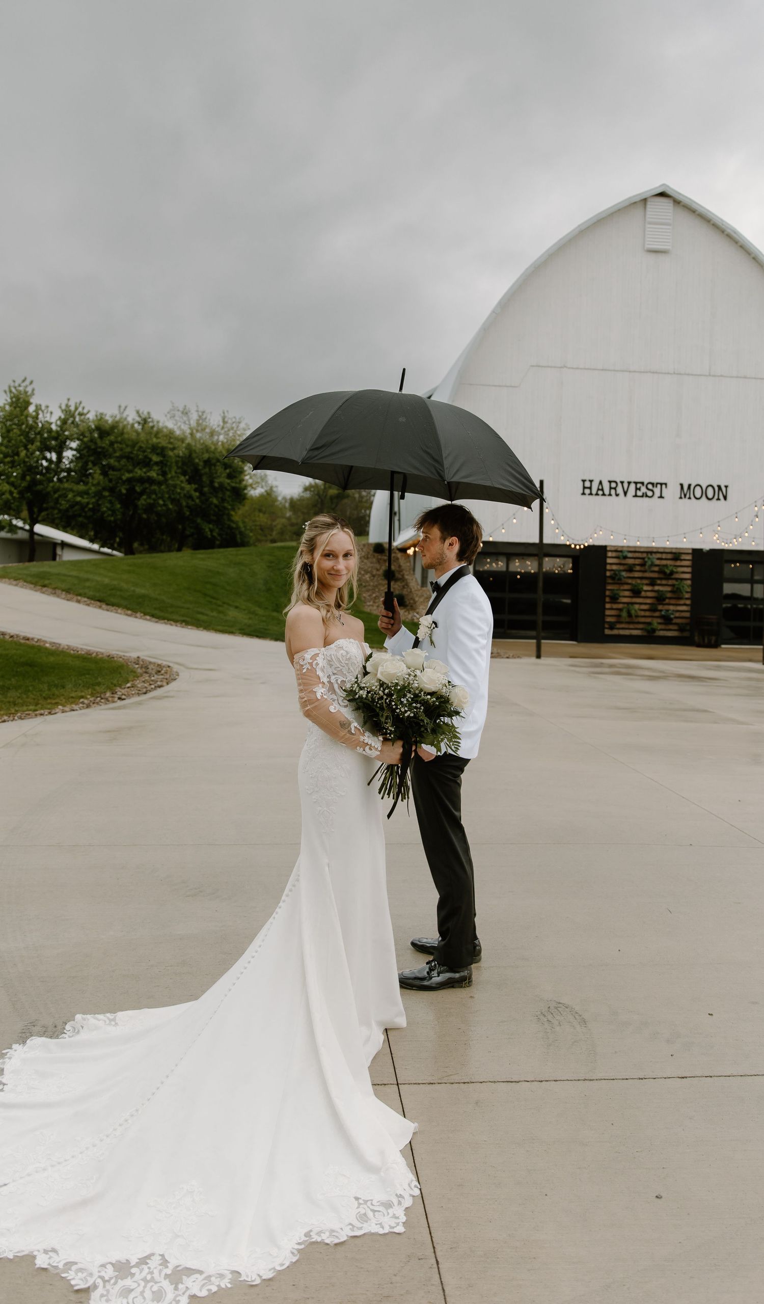 Bride and groom share umbrella outside Harvest Moon barn venue on a cloudy wedding day.