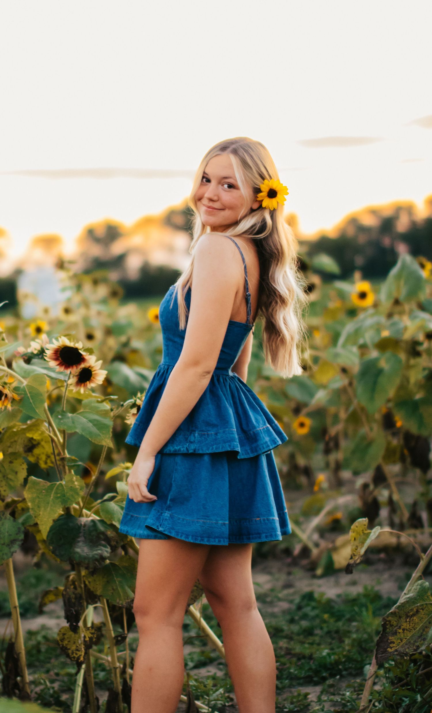 Someone in a blue ruffled dress poses playfully in a sunflower field at golden hour.