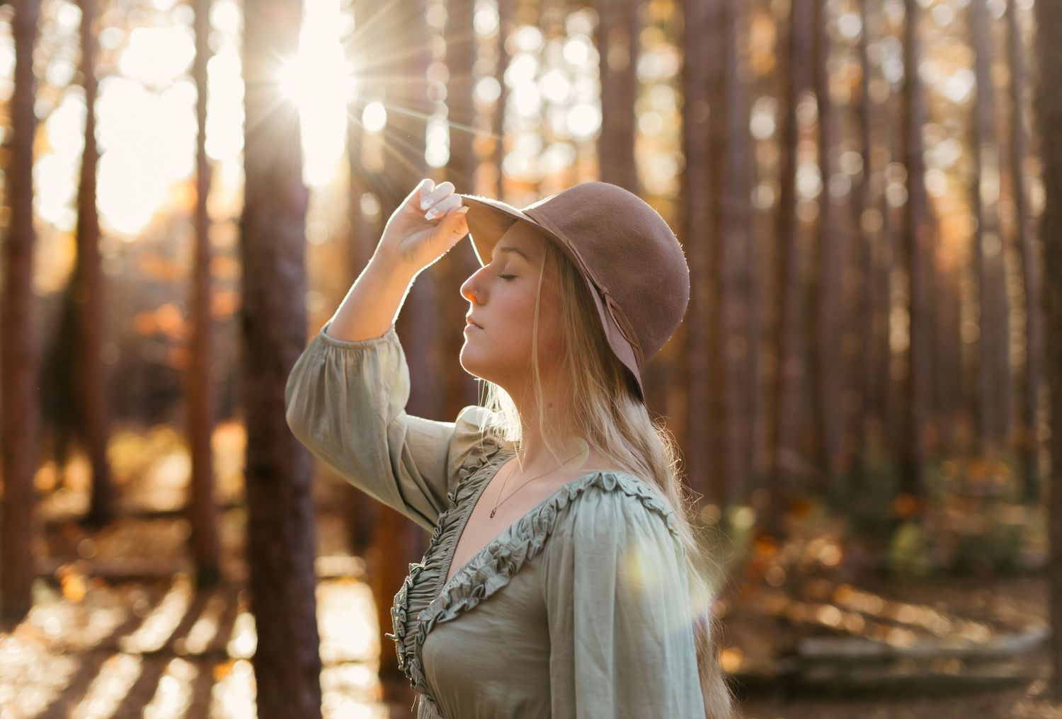 A dreamy autumn portrait of someone in a brown hat gazing at golden sunlight filtering through forest trees.
