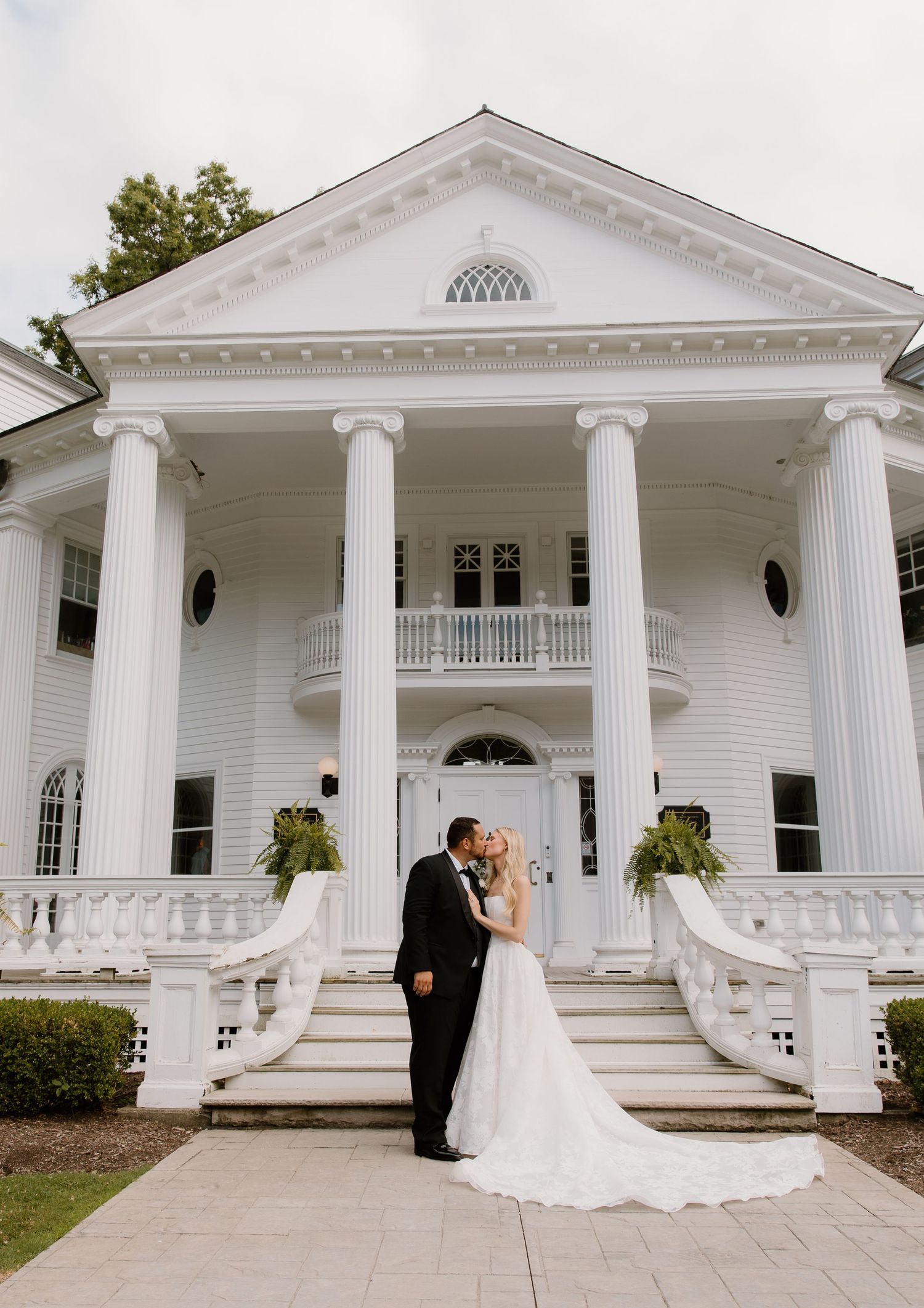 Bride and groom share romantic kiss on steps of grand white Southern mansion with tall columns.