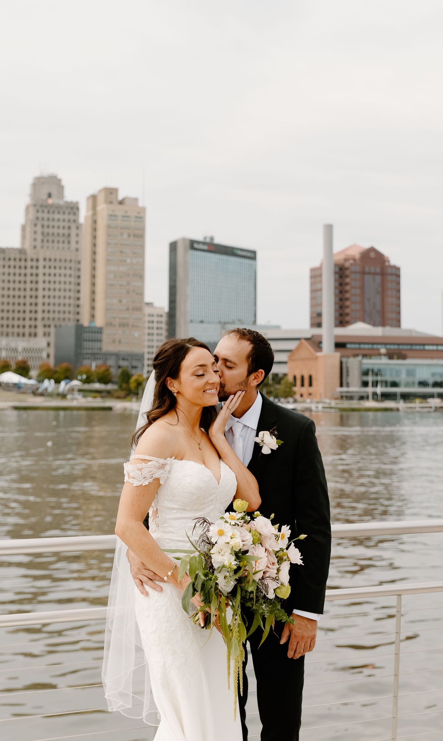 A romantic wedding couple shares intimate moments along a waterfront with a cityscape backdrop.