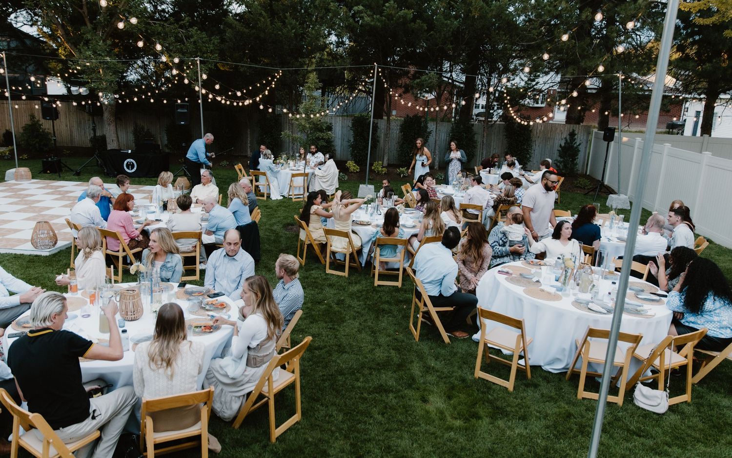 An outdoor wedding reception setup with string lights and white table settings under a tent in a garden setting.