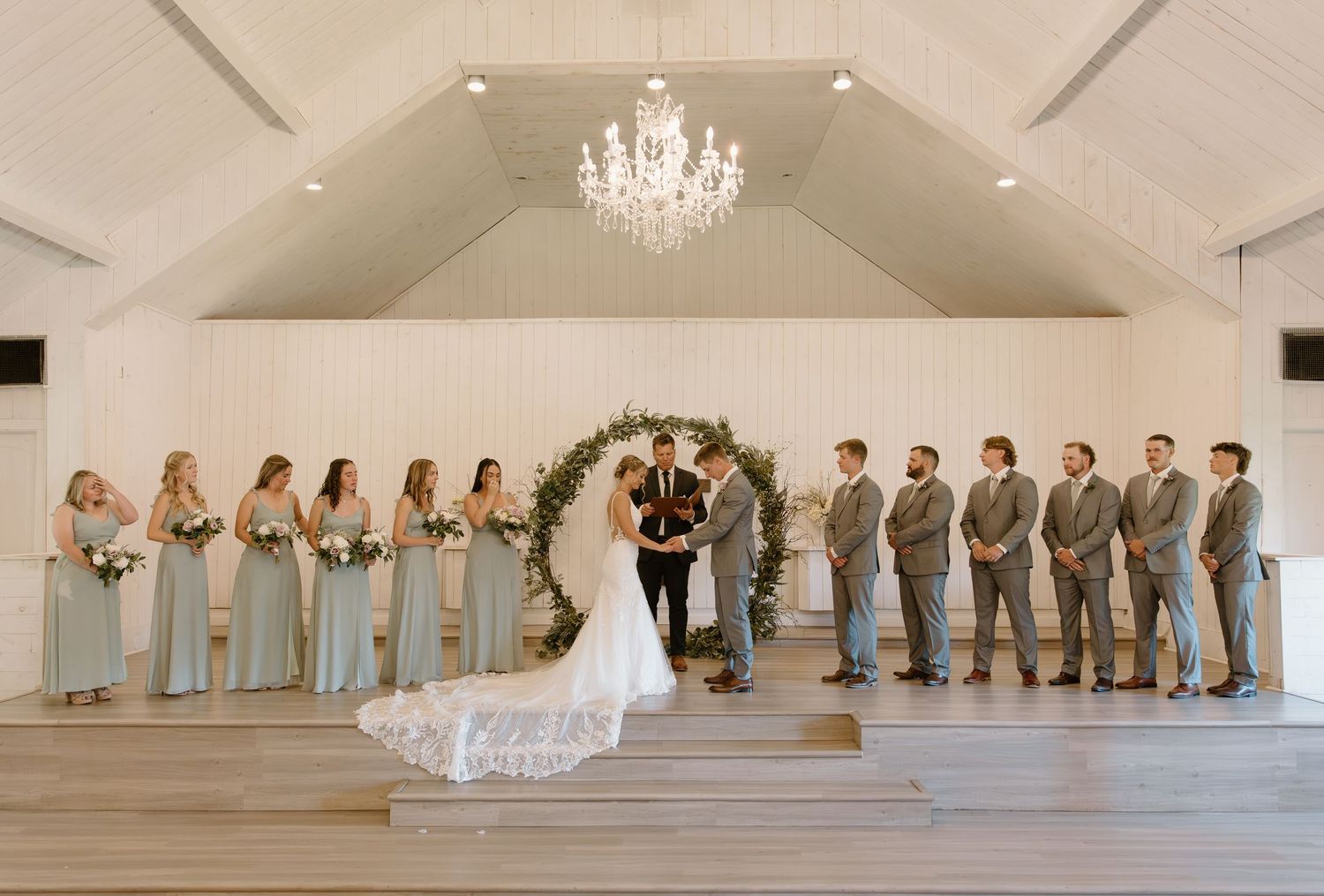 Wedding ceremony in an elegant white venue with a crystal chandelier, floral arch, and wedding party in blue and gray attire.