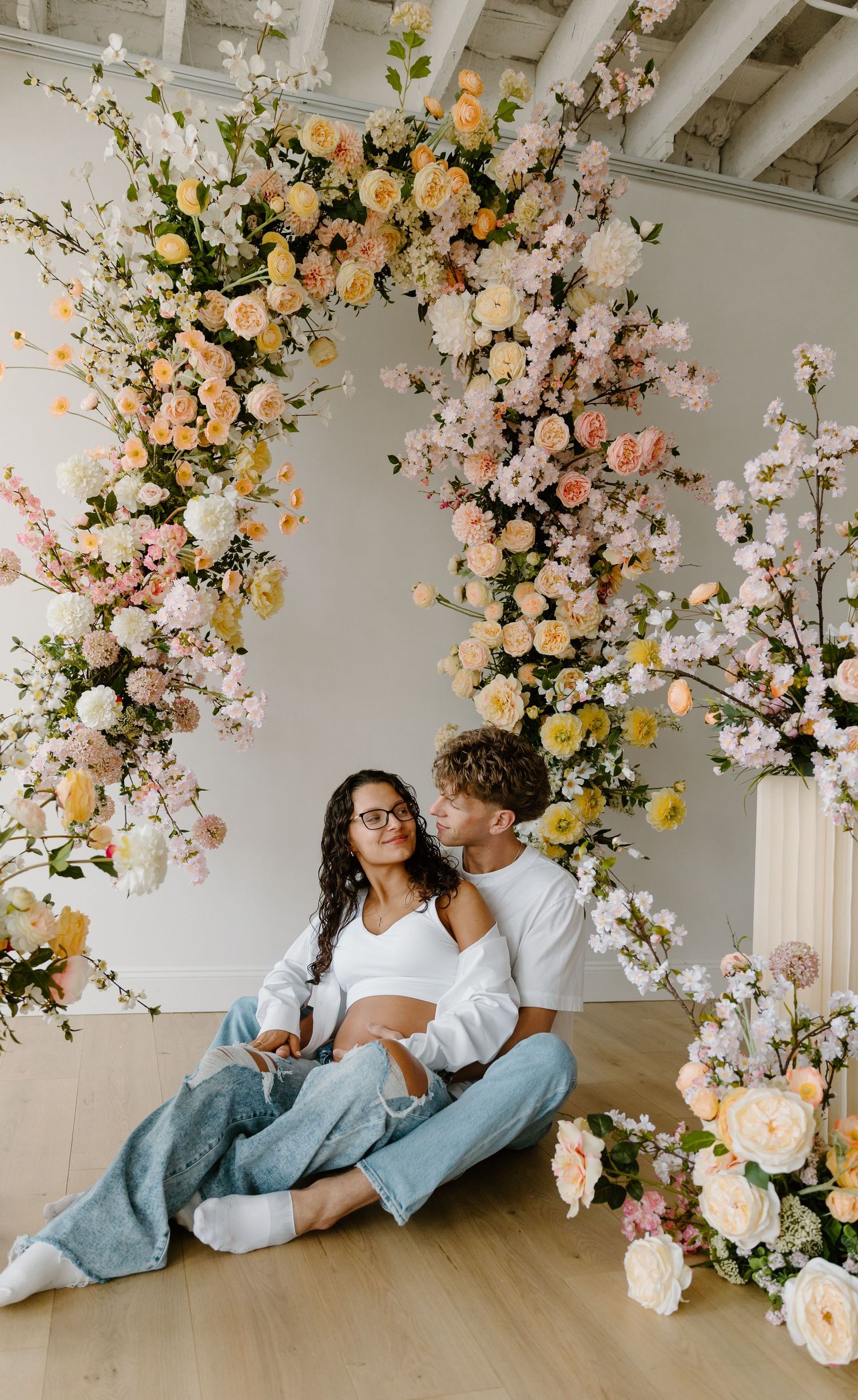 Couple sitting on floor surrounded by lush pink and peach floral arrangements in a bright studio setting.