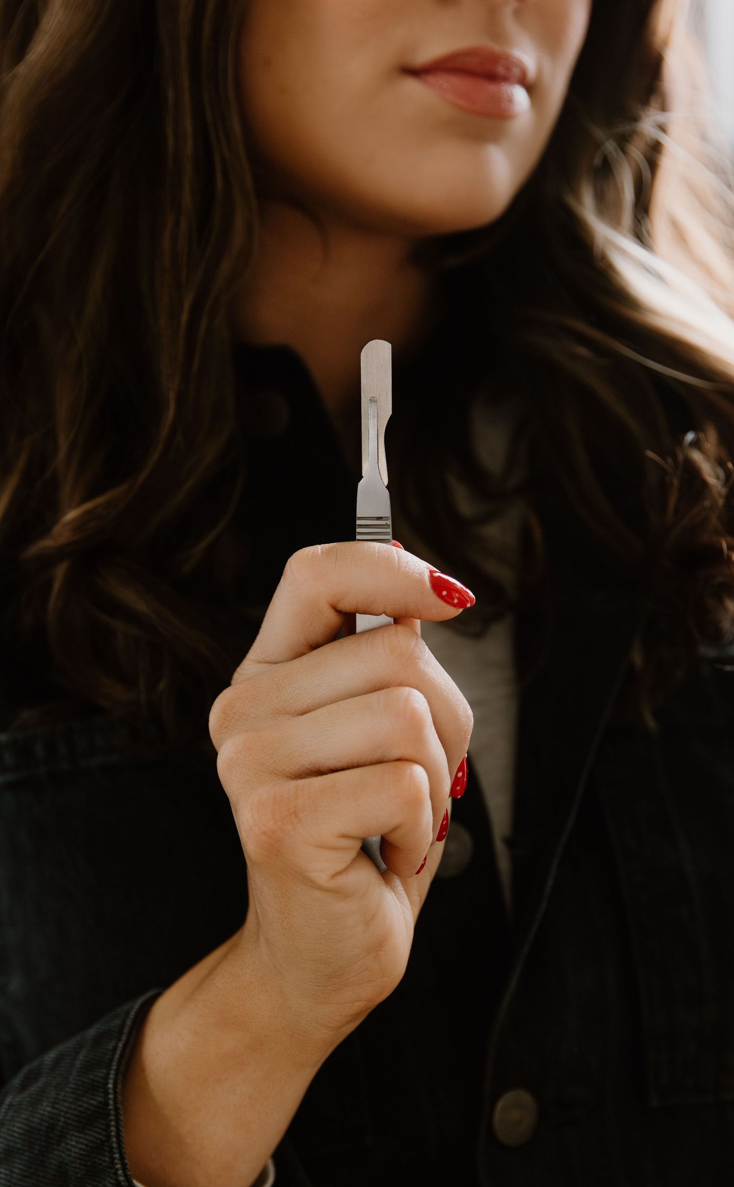 Woman with red nails smiling while holding a pregnancy test, wearing a dark jacket.