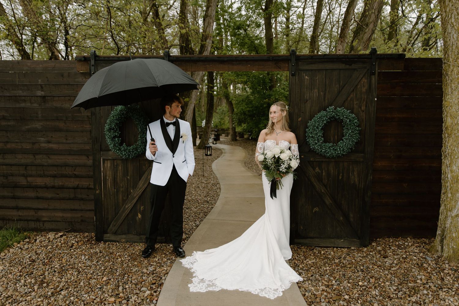 Bride in white gown and groom in tuxedo with umbrella pose before rustic barn doors with wreaths on a rainy day.
